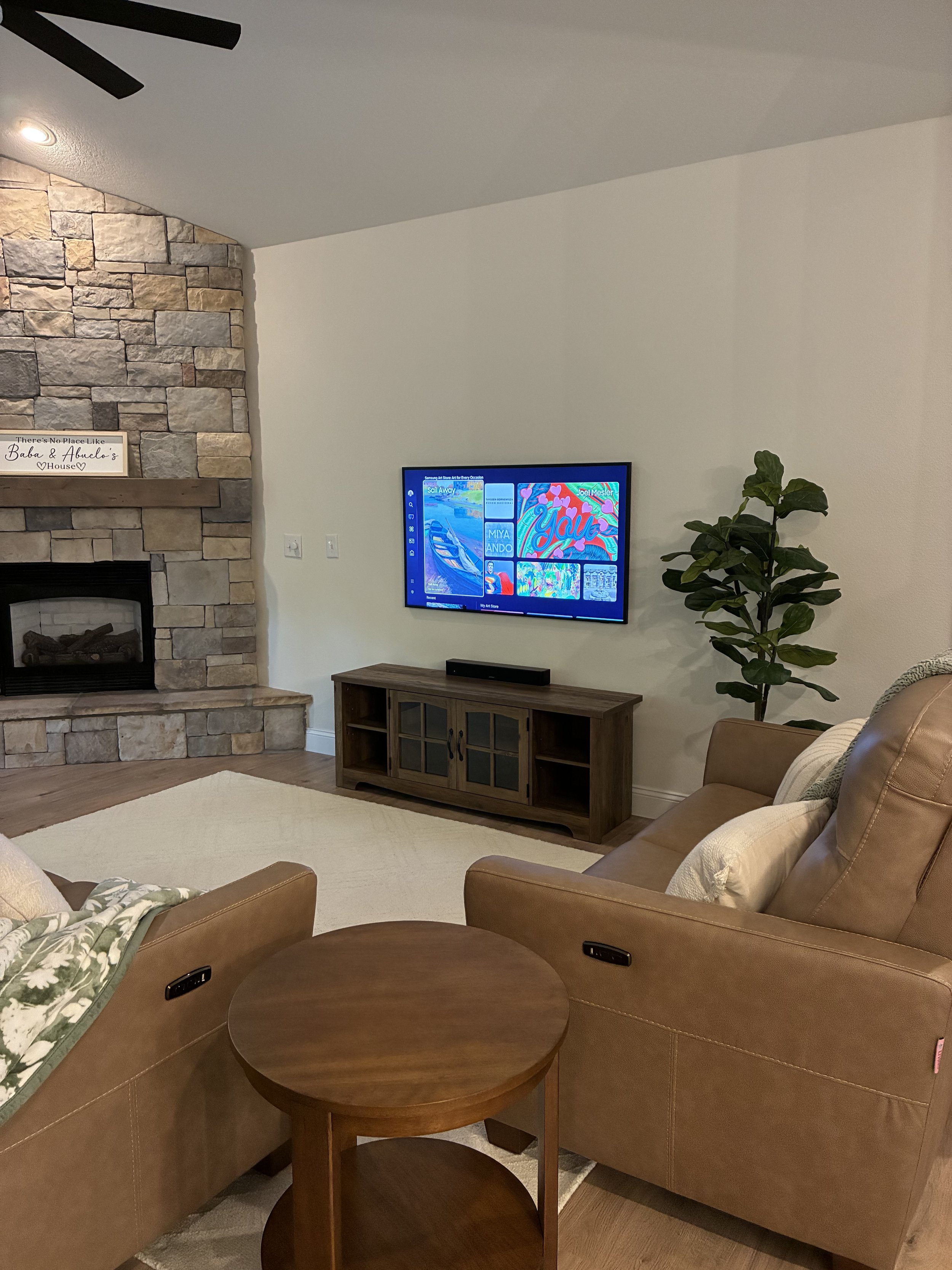 Living room with a stone fireplace on the left, a wall-mounted TV on the right, a wooden TV stand, a large houseplant, and two tan leather sofas with pillows. A small round wooden side table is between the sofas.