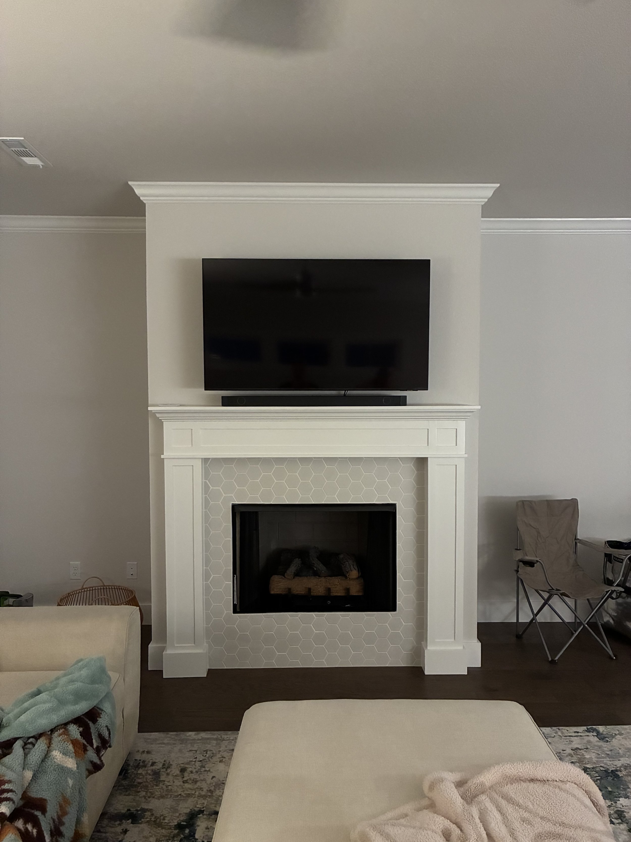 Living room with a white fireplace and a flat-screen TV mounted above it, beige sofa in the foreground, a soft blanket on the sofa, and a folding chair to the right.