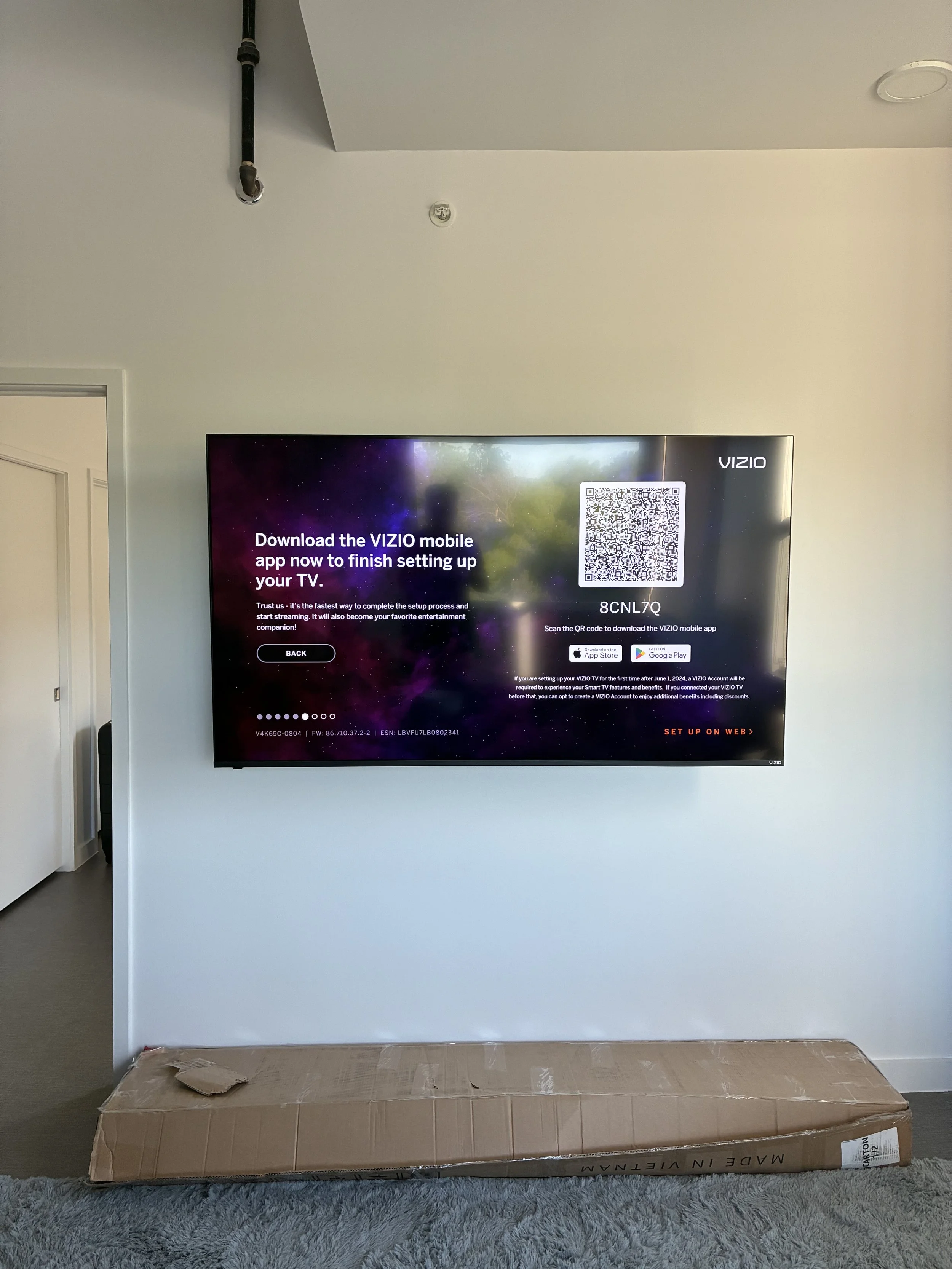Flat screen TV mounted on a white wall displaying setup instructions with a QR code and app download options. A cardboard box is on the floor below the TV.