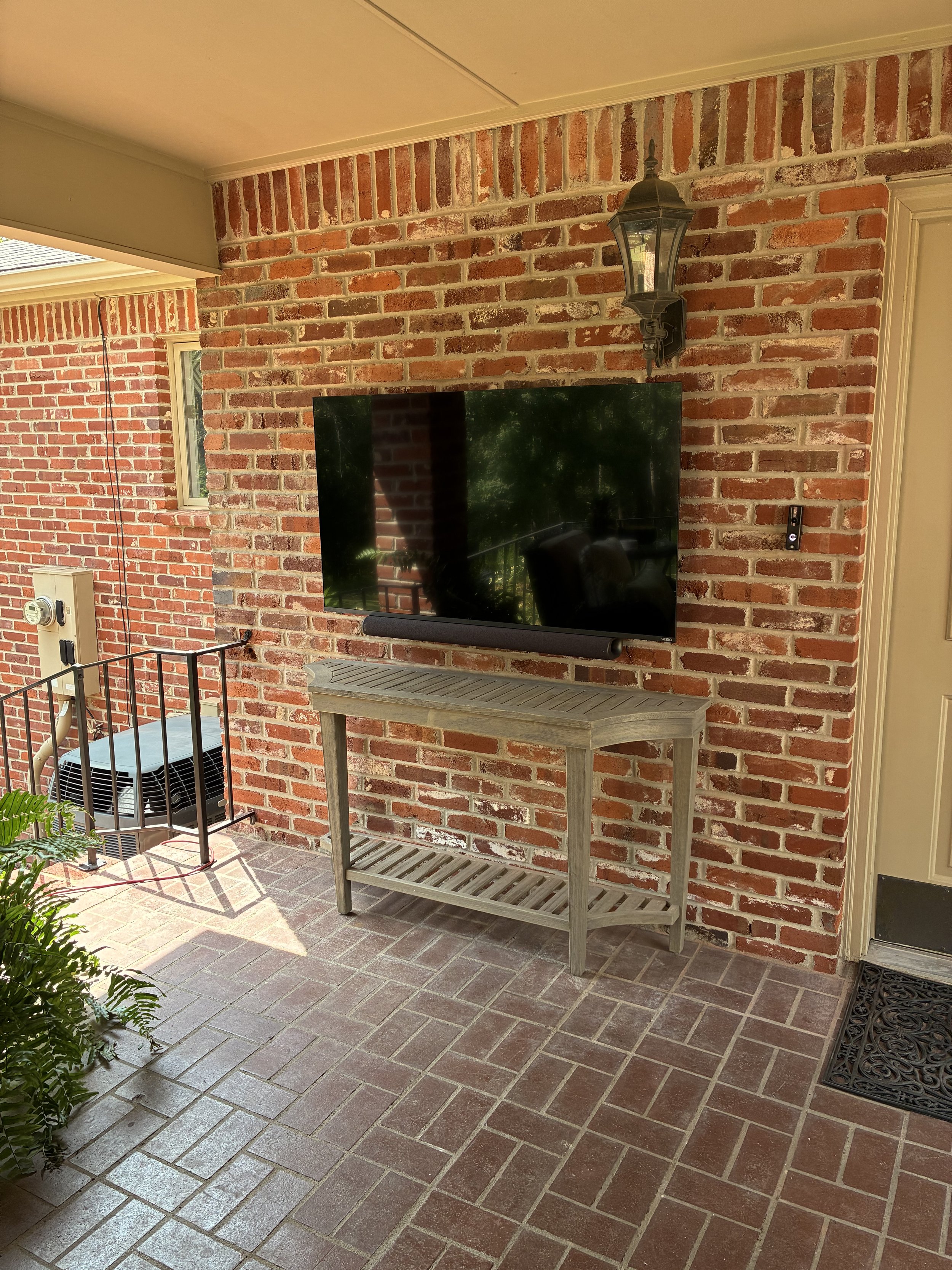 Outdoor porch area with brick wall, a large flat-screen TV mounted above a wooden table, and a decorative outdoor lantern light fixture.