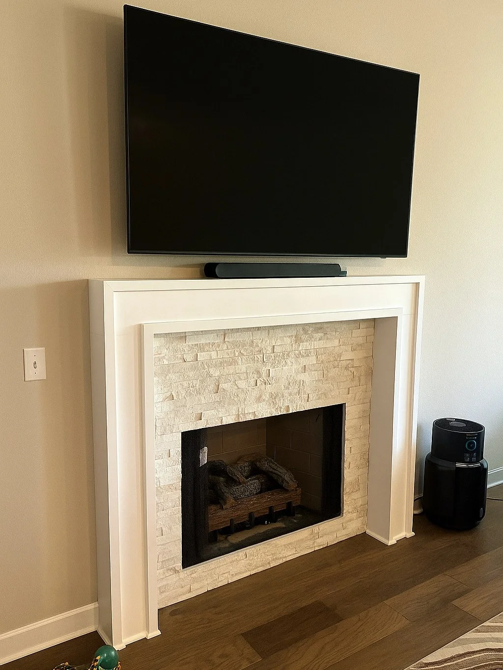 Flat-screen TV on a white fireplace mantel with a stone interior, next to a black air purifier on the wooden floor.