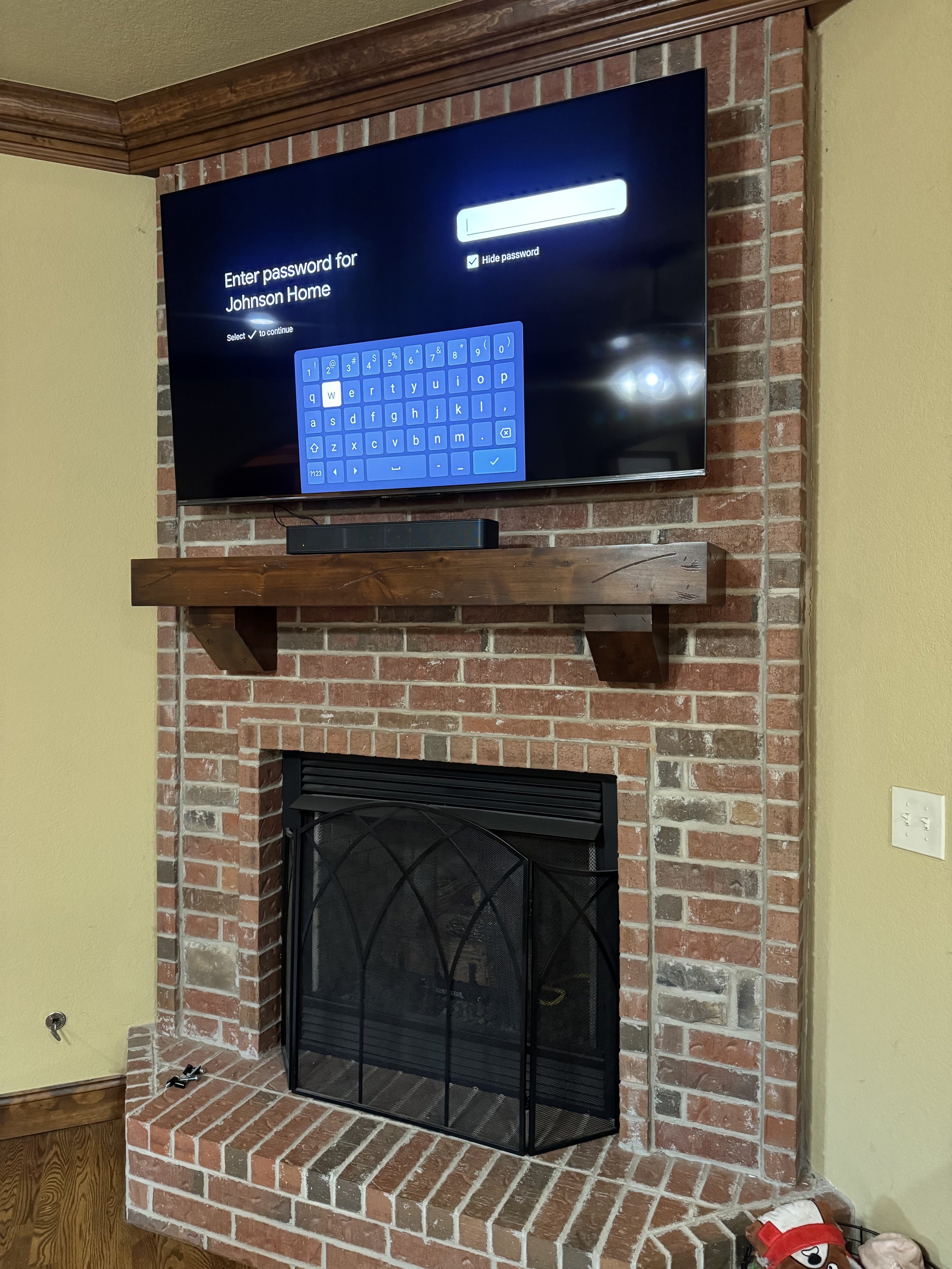 TV mounted above a brick fireplace with a wooden mantel. The TV screen shows a prompt to enter a password for 'Johnson Home'.
