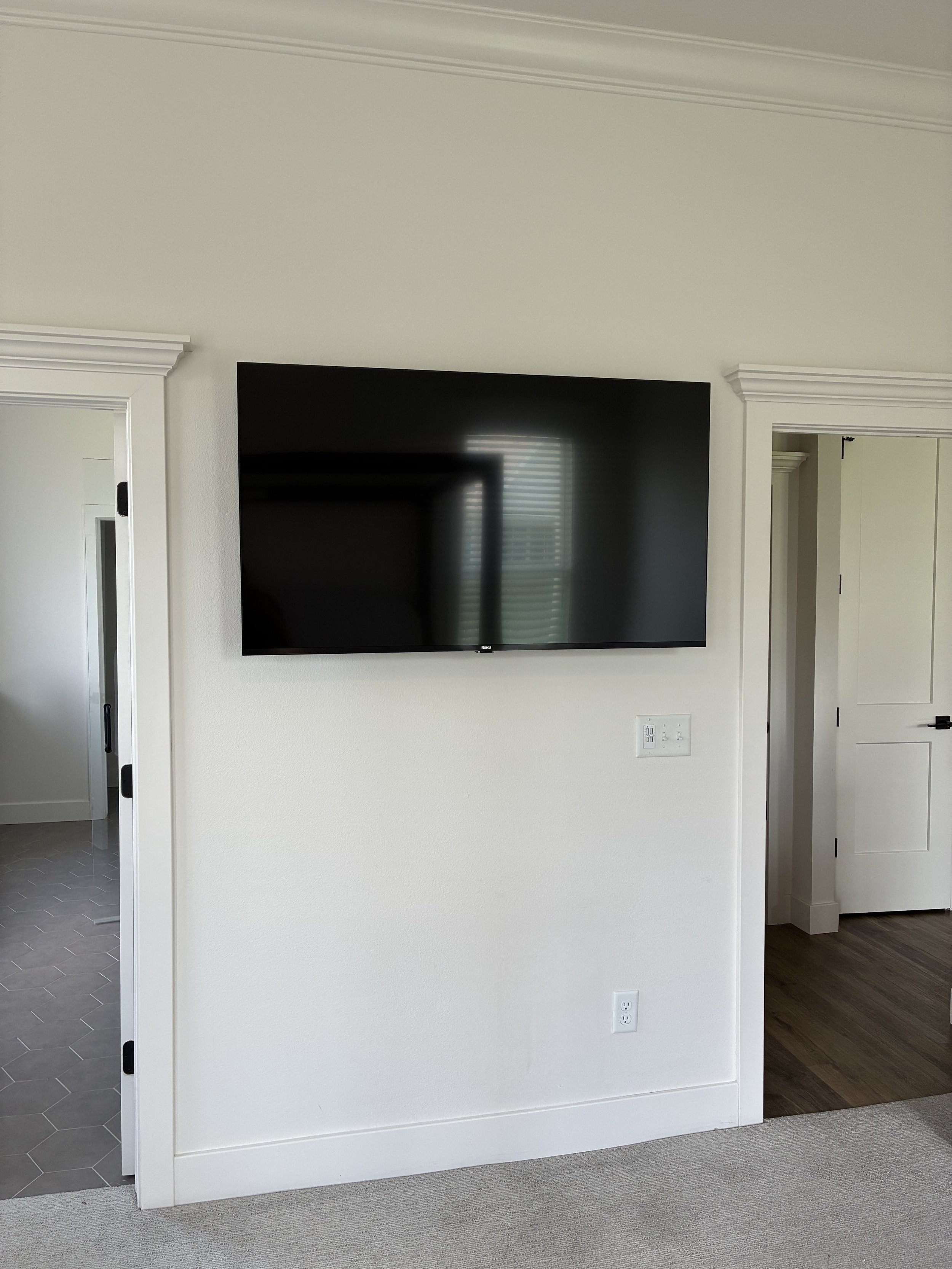 A flat-screen television mounted on a white wall in a modern living room, with a reflection of a window with blinds visible on the screen.