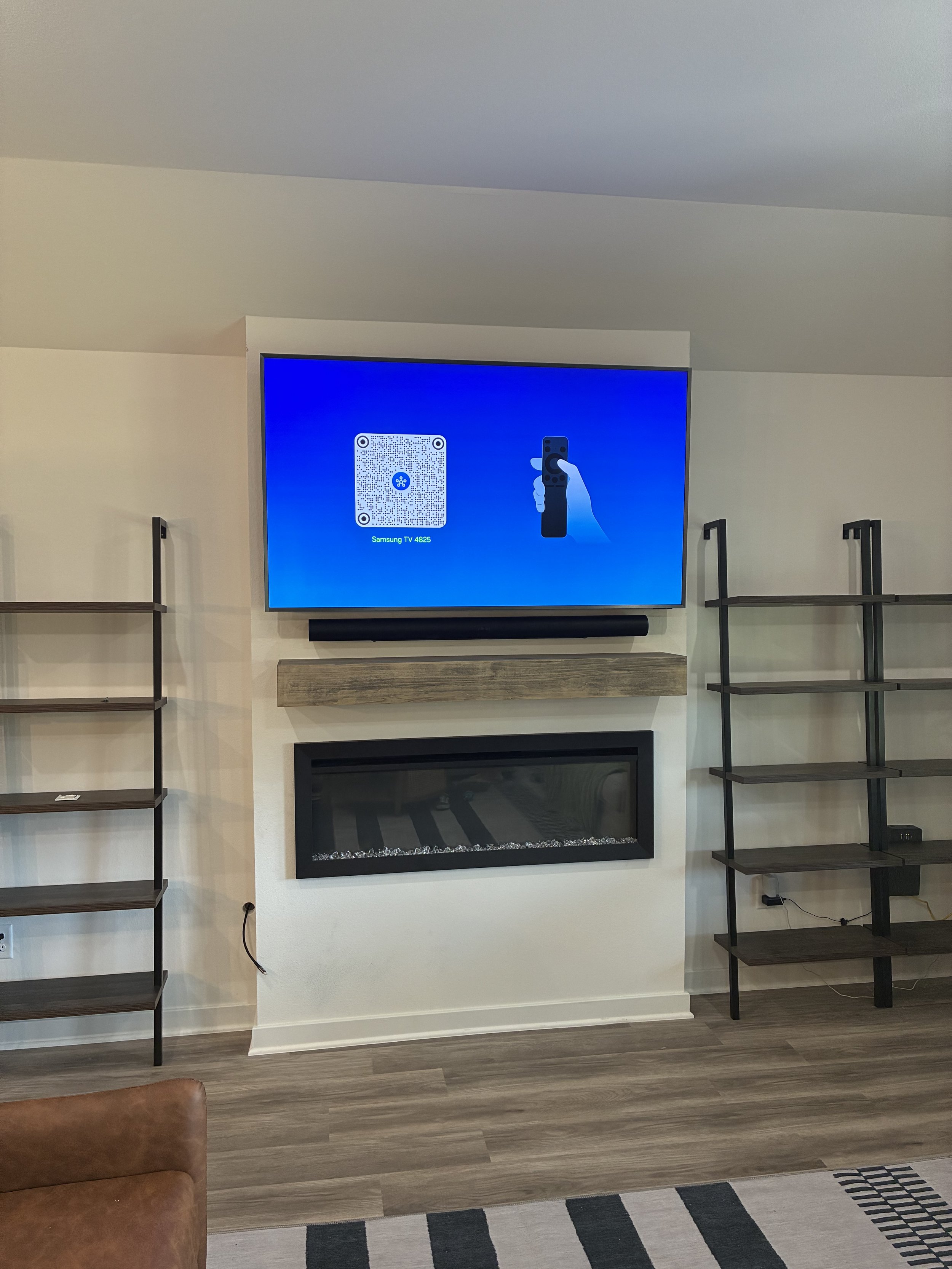 Living room with a mounted TV screen displaying a QR code and a remote control, a modern fireplace, and empty black shelving units on each side.