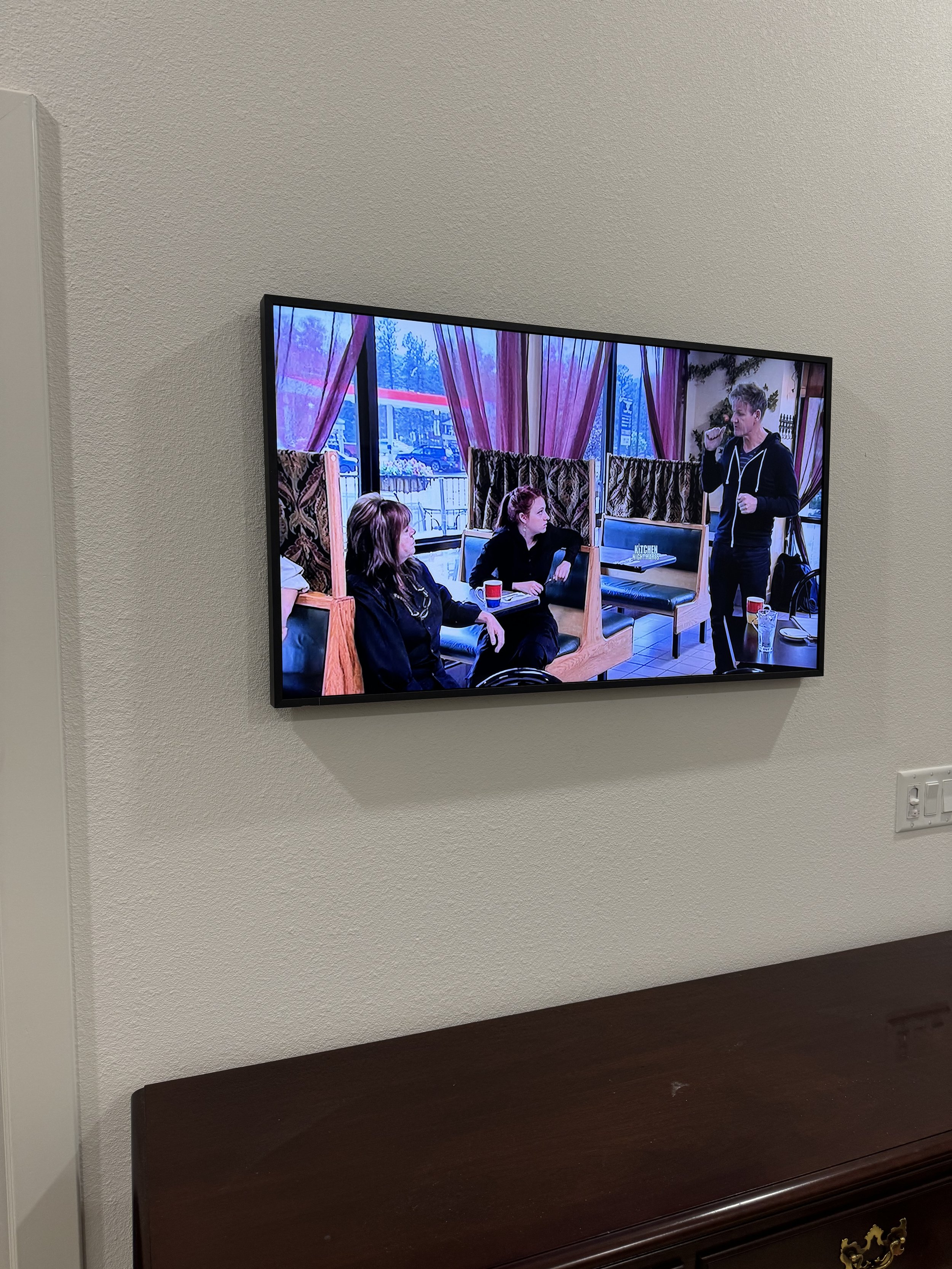 TV screen displaying three women seated at a wooden booth table in a restaurant or cafe, with a man standing nearby, talking into a microphone or headset, during daytime.