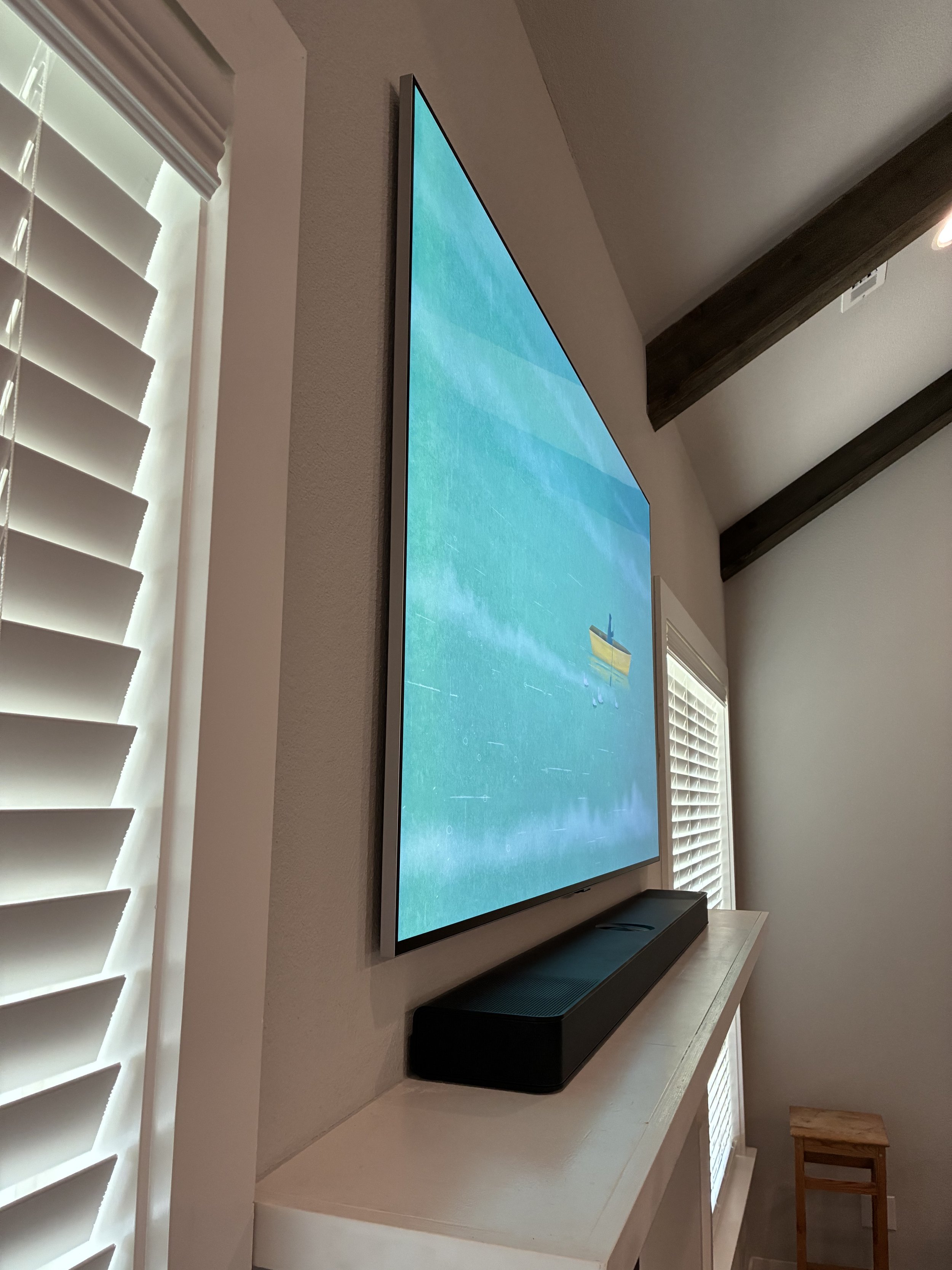 A wall-mounted flat-screen TV displaying a scene of a boat on calm water, with a black soundbar placed below it on a white shelf in a room with wooden ceiling beams and window shutters.