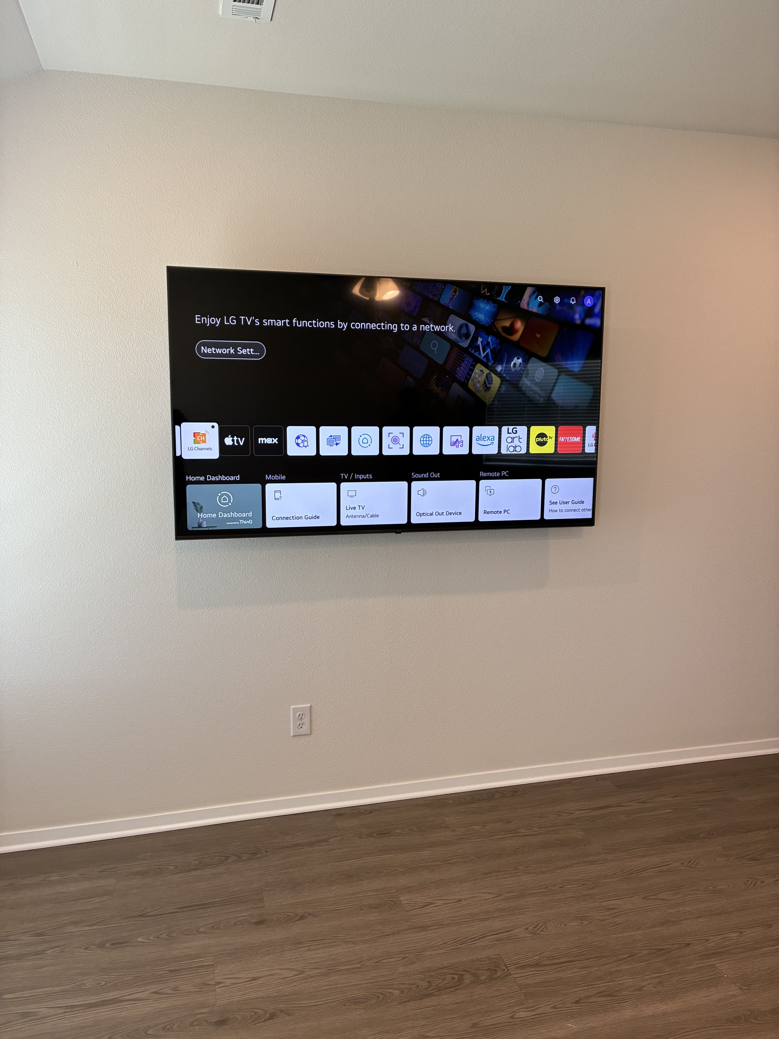 A wall-mounted flat screen TV displaying setup menu, located on a plain beige wall above a wooden floor, with an electrical outlet below.