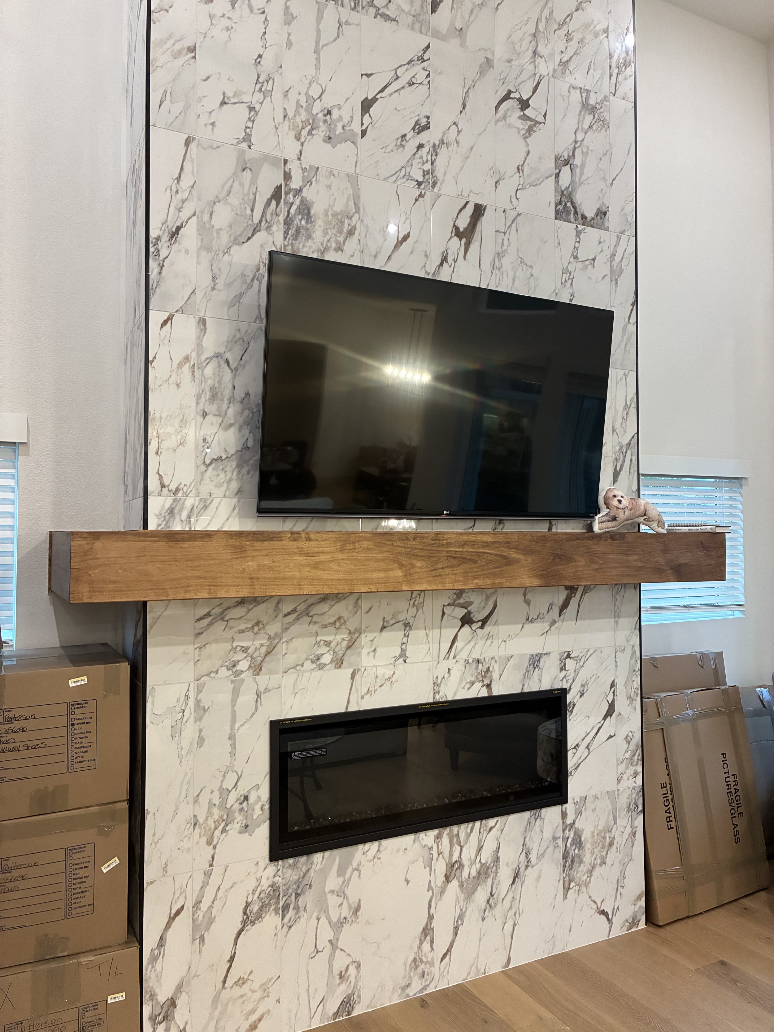 A television mounted on a marble fireplace wall with a wooden mantel. There are stacked boxes on the floor and windows with blinds on either side of the fireplace. A decorative animal figurine is on the mantel.