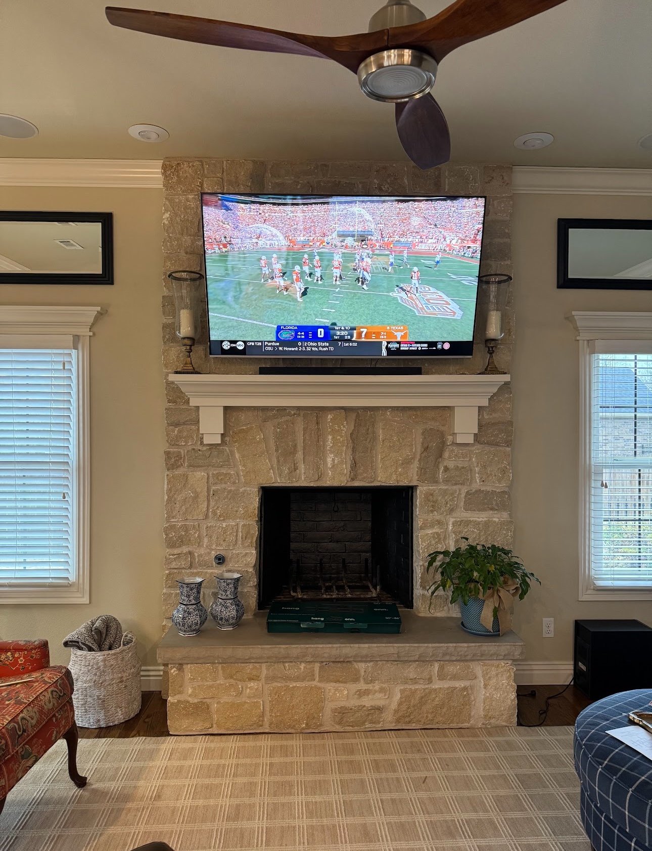Living room with a stone fireplace, a wall-mounted TV showing a football game, windows with white blinds, a ceiling fan, and decorative vases and a plant on the fireplace mantle.