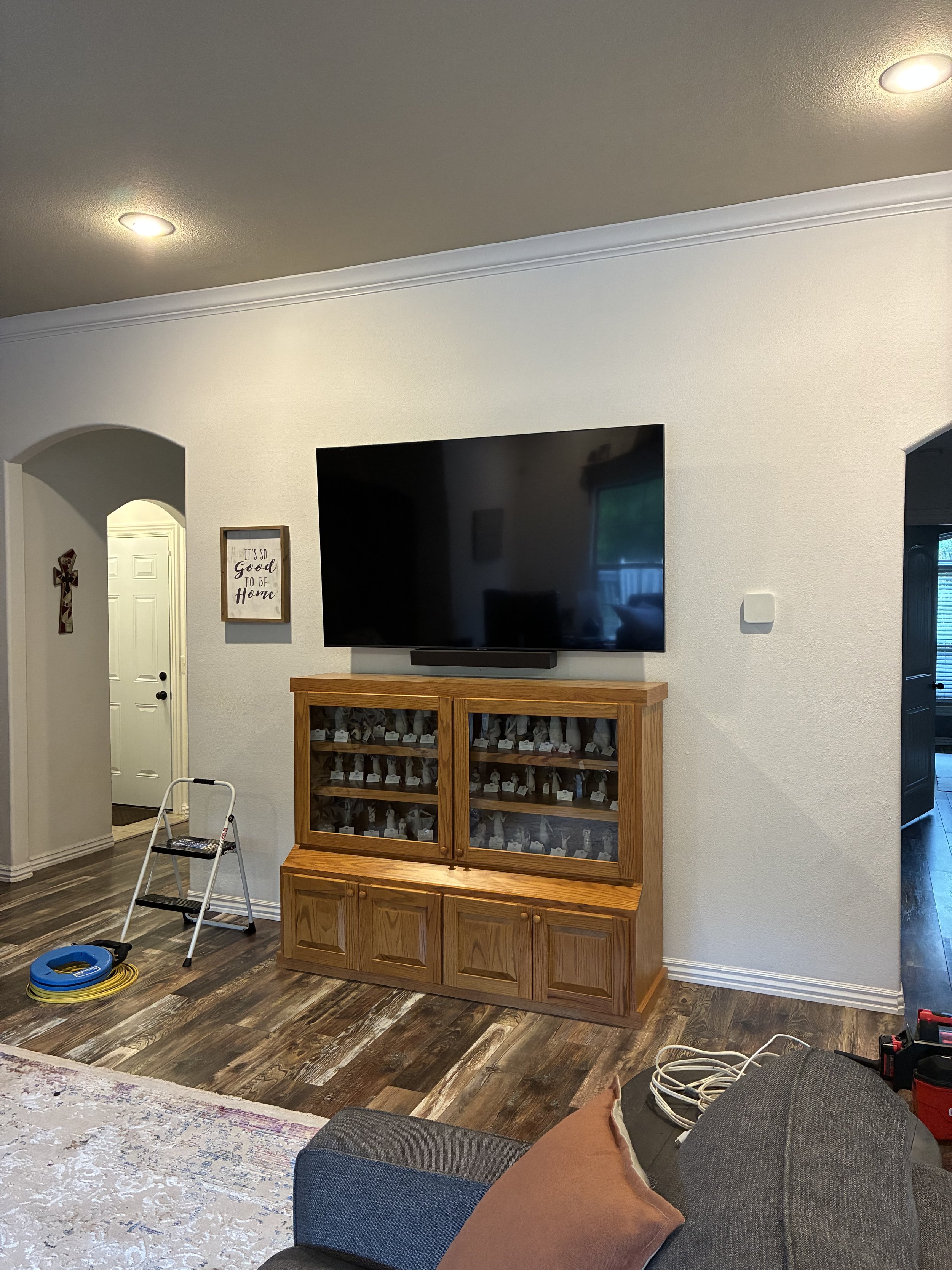 Living room with a large flat-screen TV mounted on a white wall above a wooden cabinet with glass doors. To the left of the TV is a framed sign that reads 'It's so good to be home.' There's a stepped ladder and a rolled-up extension cord on the floor