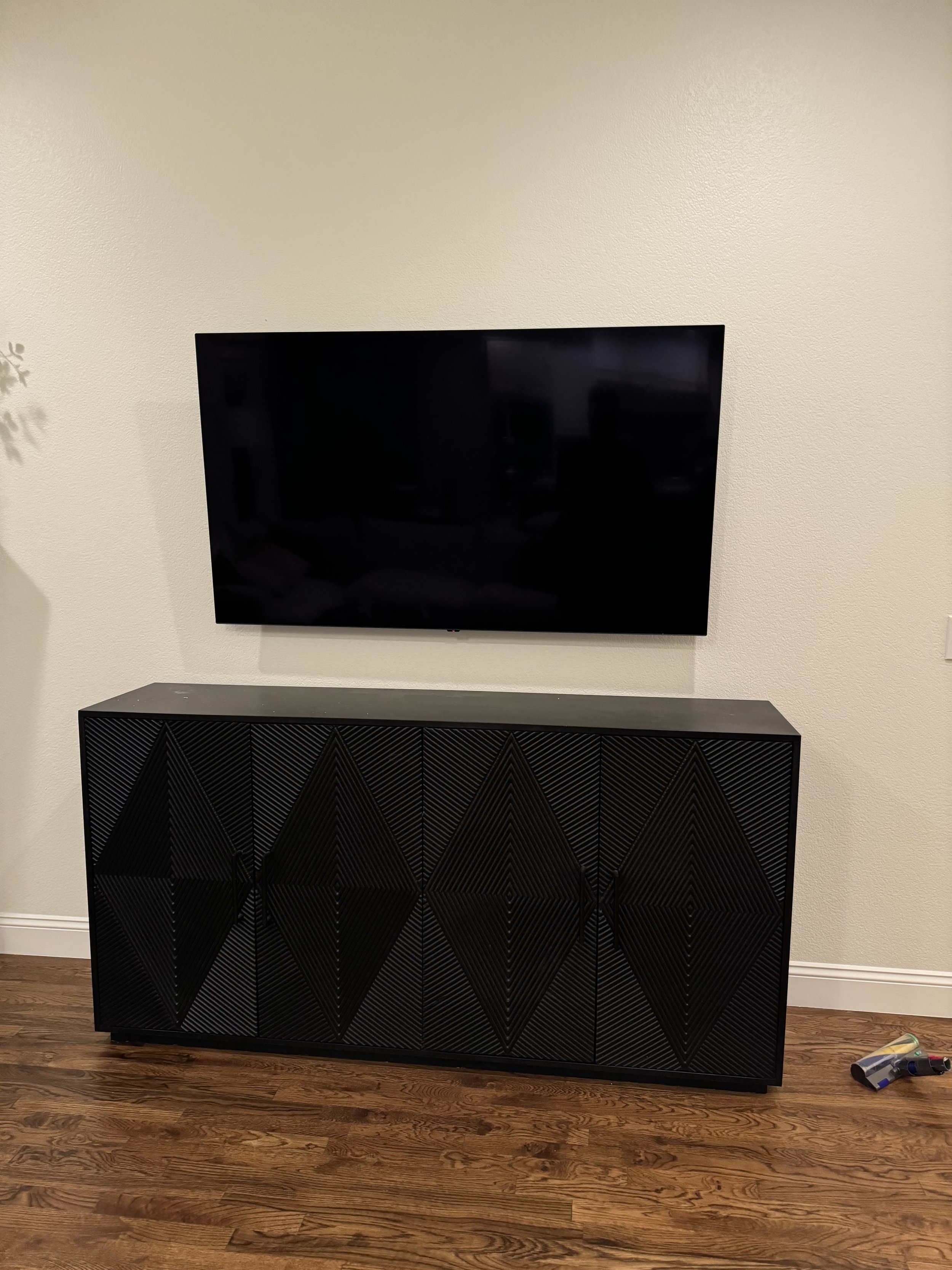 A flat-screen TV mounted on a wall above a black textured cabinet with diagonal patterns, against a beige wall, with hardwood floor and a small toy on the floor.