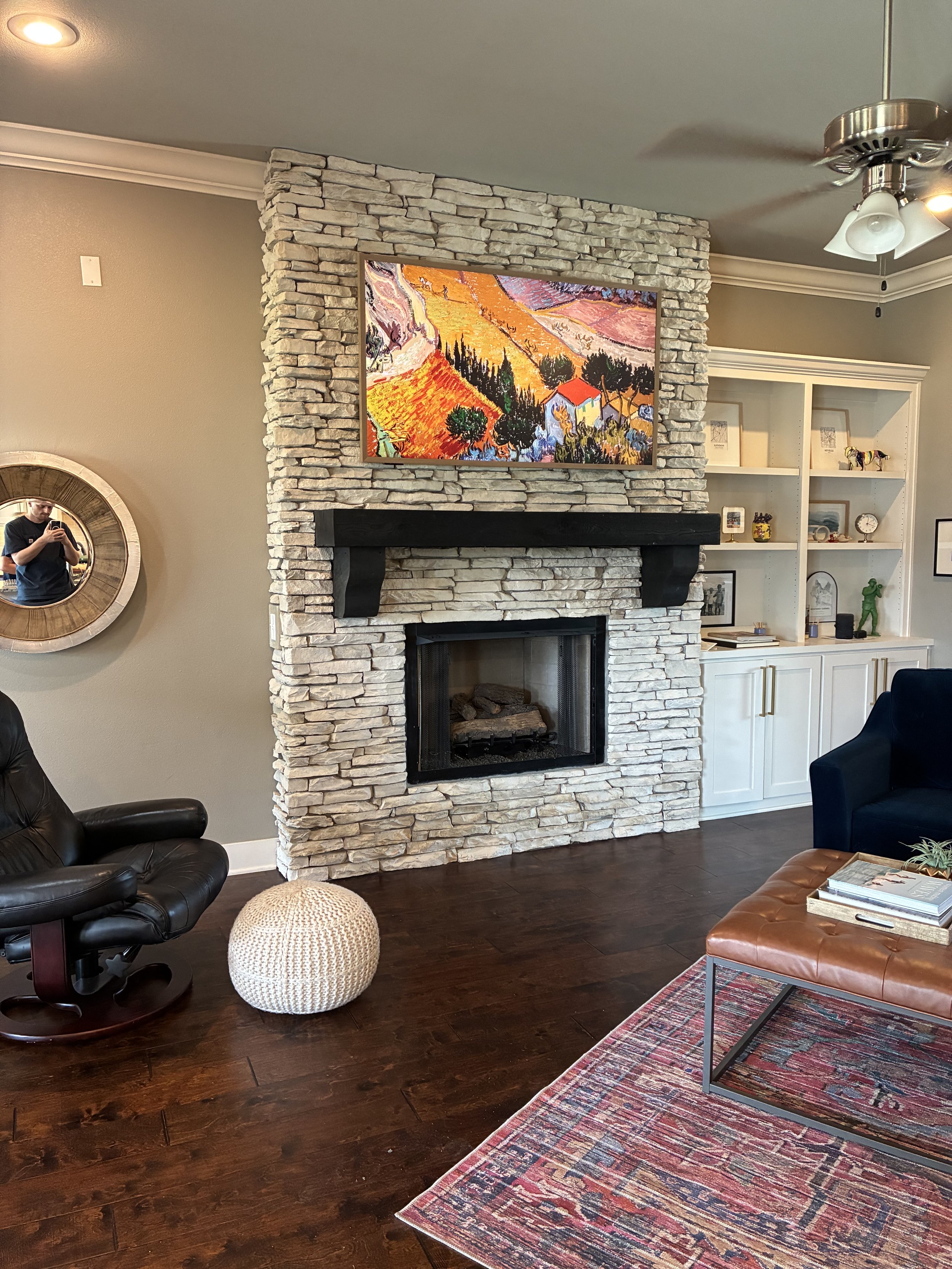 Living room with a stone fireplace, a mounted flat-screen TV displaying colorful landscape art, a black armchair, a knitted pouf, a brown leather ottoman, and a white built-in shelf with decorations.