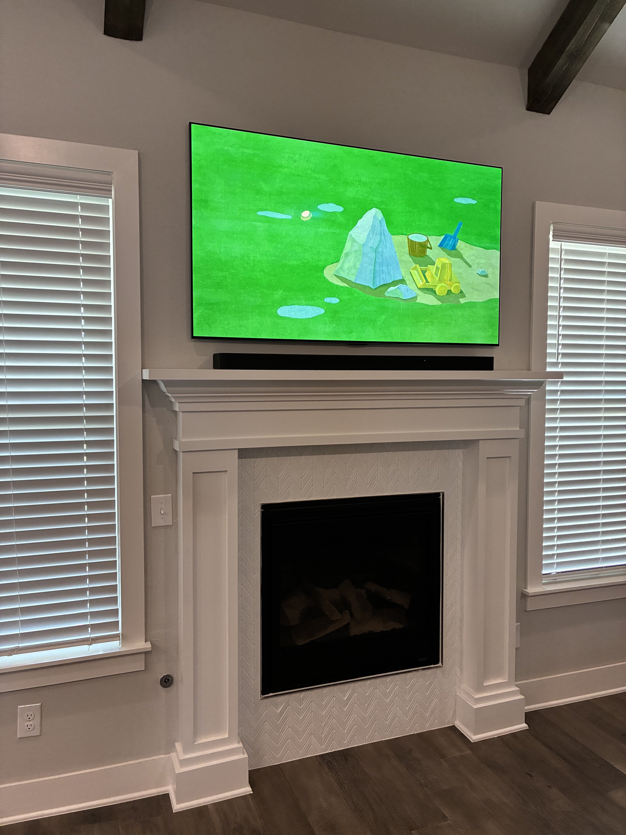 Television mounted above a white fireplace mantle showing a cartoon beach scene with a sandcastle, water puddles, a bucket, a shovel, and a toy truck.
