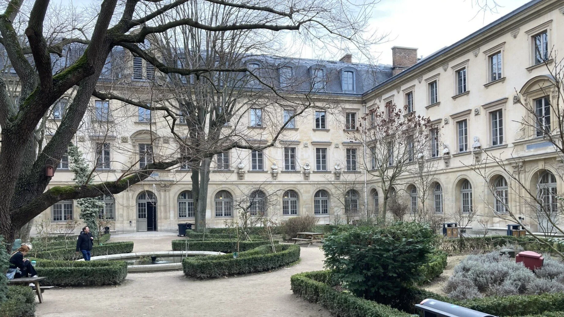 A courtyard with trees, bushes, benches, and a fountain, surrounded by a four-story historic building with ornate windows and bust sculptures on the facade.