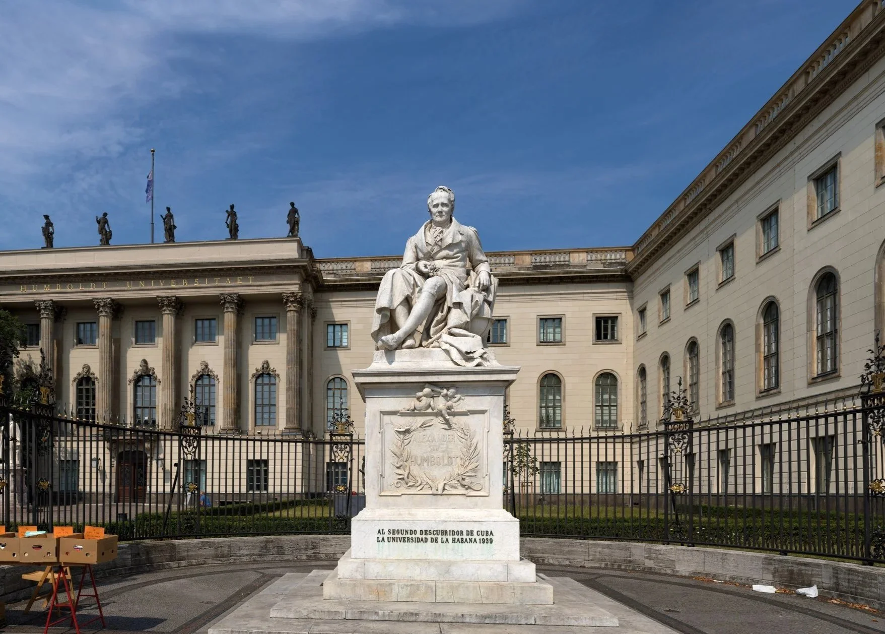 Statue of a seated man in front of a historic building with Corinthian columns and Greek statues on the roof, with a fence and a few boxes on the sidewalk.