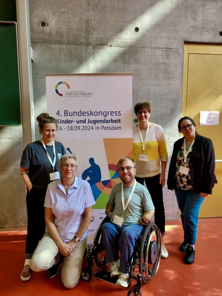 Gruppe von fünf Menschen, darunter eine Person im Rollstuhl, vor einem Banner für den 4. Bundeskongress Kinder- und Jugendarbeit in Potsdam, mit sichtbarem Datum 16. bis 18. September 2024.