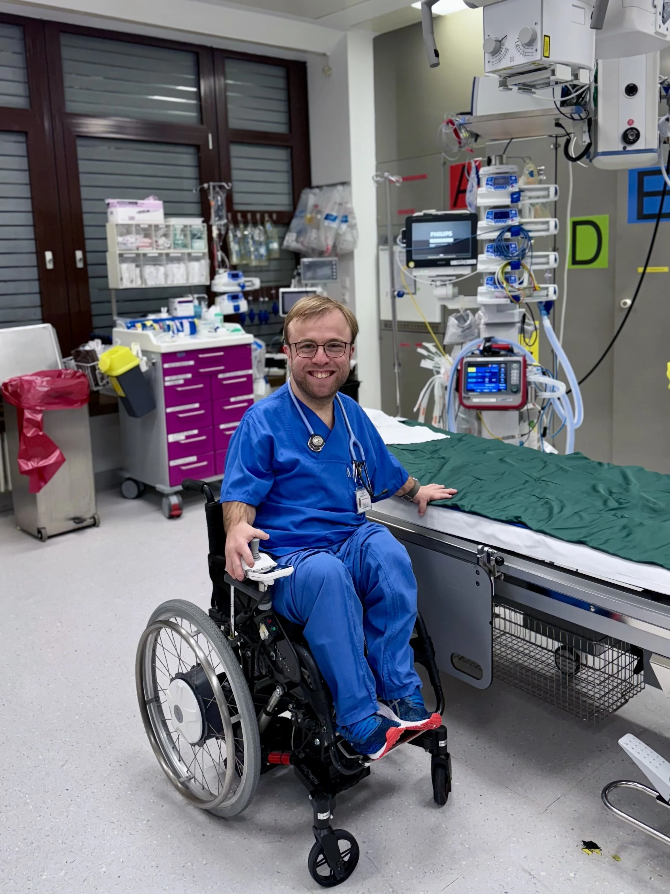 Ein Mann im blauen Krankenhauskittel sitzt im Rollstuhl in einem medizinischen Behandlungsraum, umgeben von medizinischer Ausrüstung und Geräten.