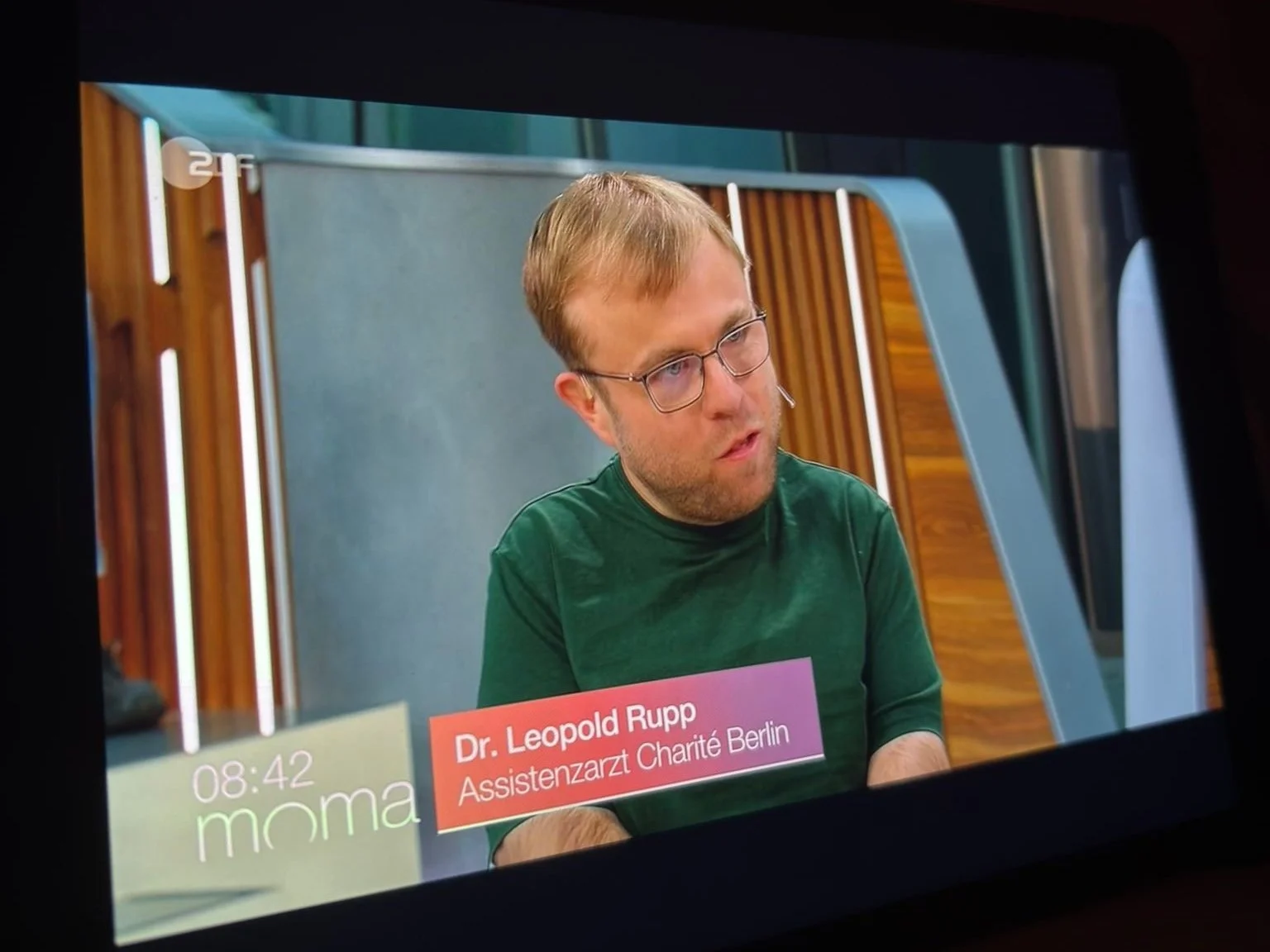 Ein Mann mit Brille und einem grünen Pullover sitzt an einem Tisch. Im Bild ist eine Bildunterschrift mit dem Namen Dr. Leopold Rupp, Assistenzarzt bei Charité Berlin, zu sehen.