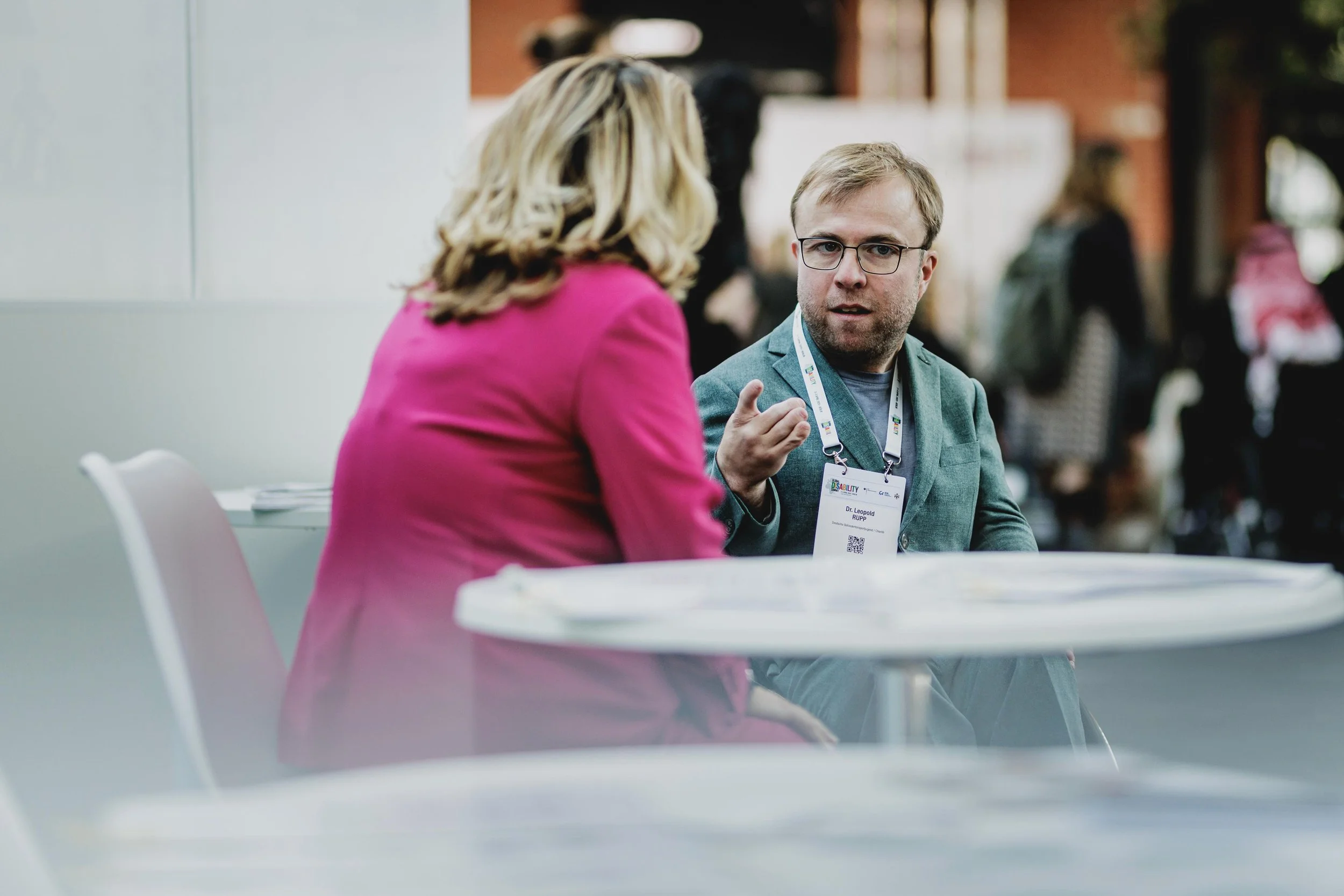 Zwei Menschen sprechen bei einem Treffen, der Mann trägt eine Namensschild, die Frau trägt einen pinken Blazer.