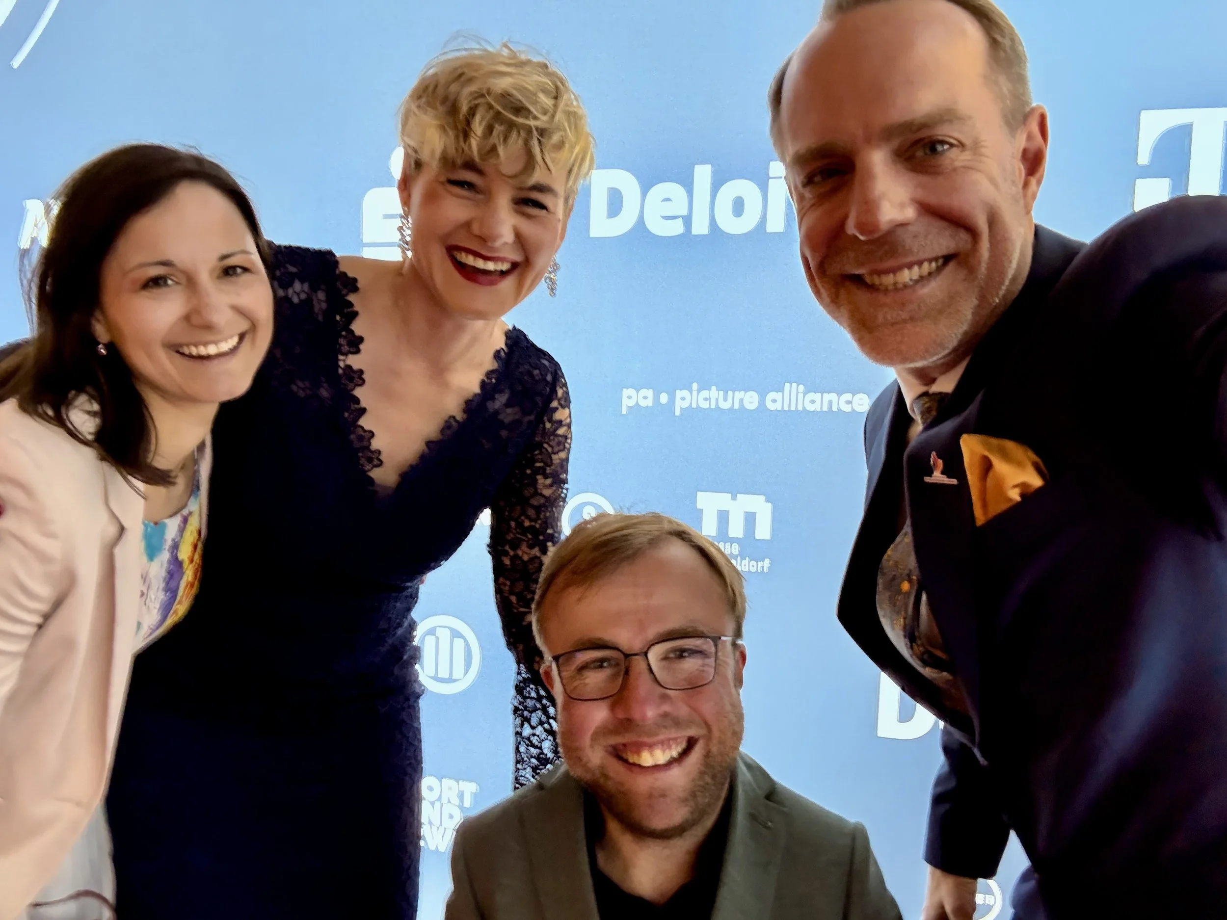 Fünf Menschen, vier Frauen und ein Mann, posieren lachend für eine Gruppefoto vor einem blauen Hintergrund mit logos.