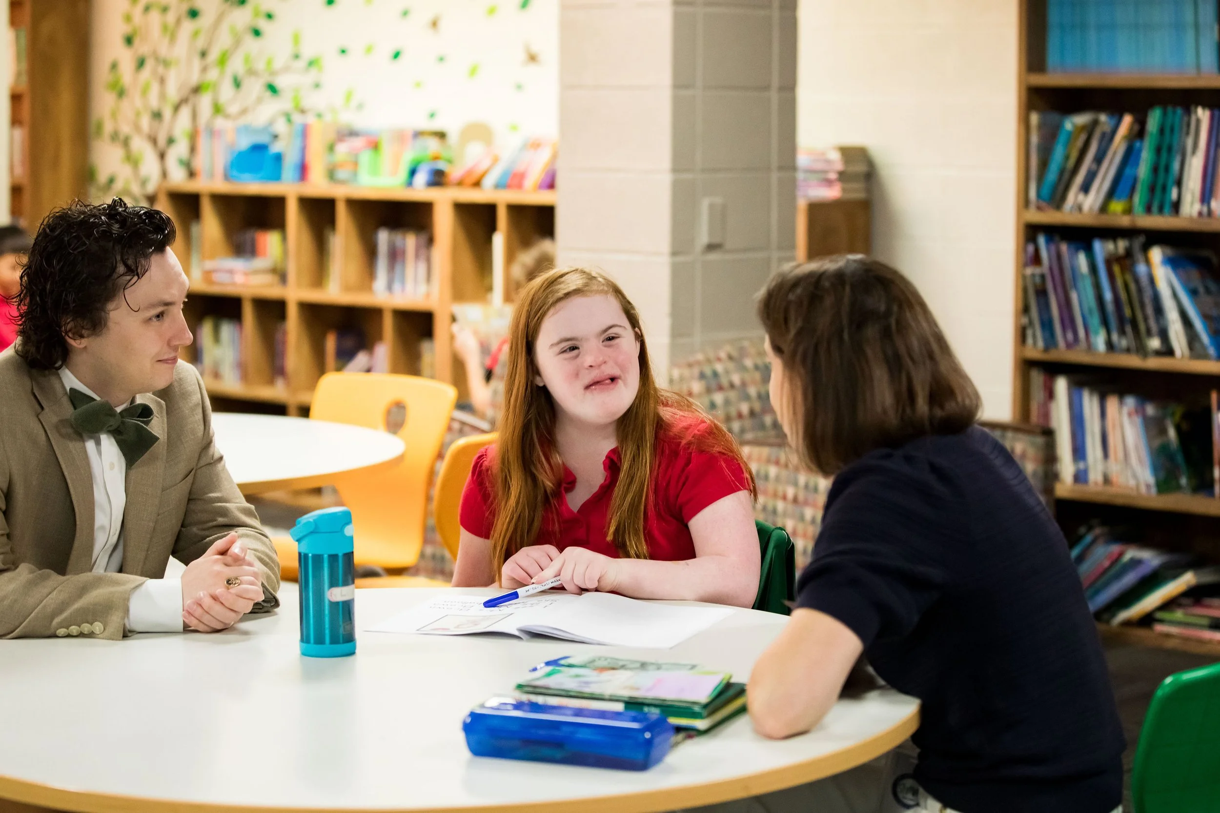 A group of people sitting around a table in a library, engaged in conversation. A man in a beige suit and green bowtie sits on the left, a girl with long red hair wearing a red shirt is in the middle, and a boy in a navy blue shirt is on the right. Bookshelves are in the background.