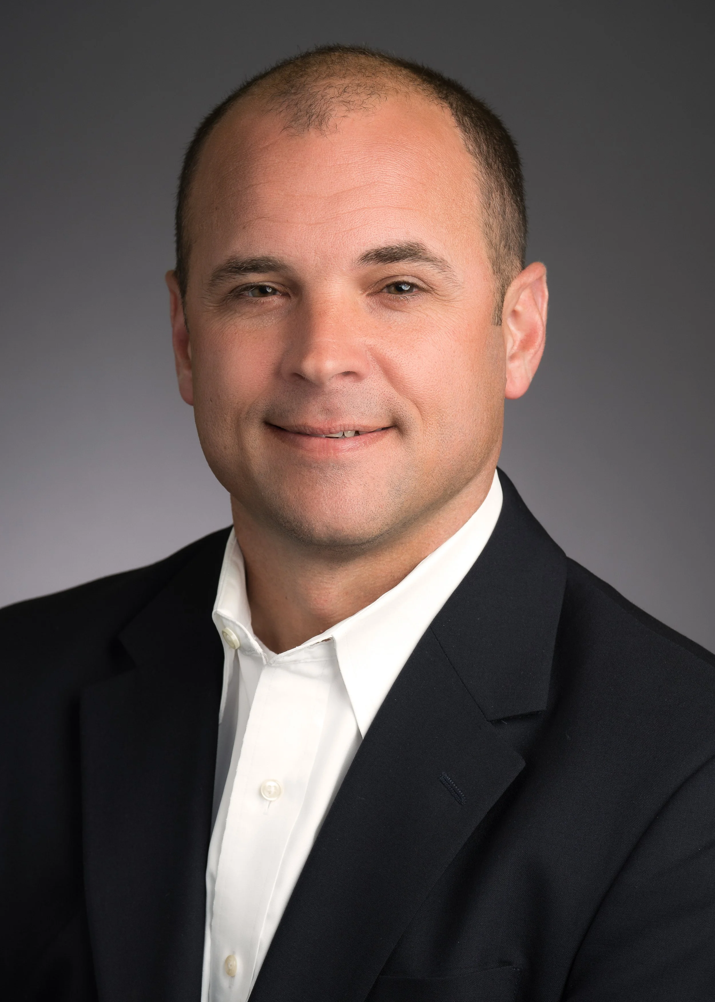 Professional headshot of a man wearing a black suit and white shirt, smiling slightly, set against a gray background.