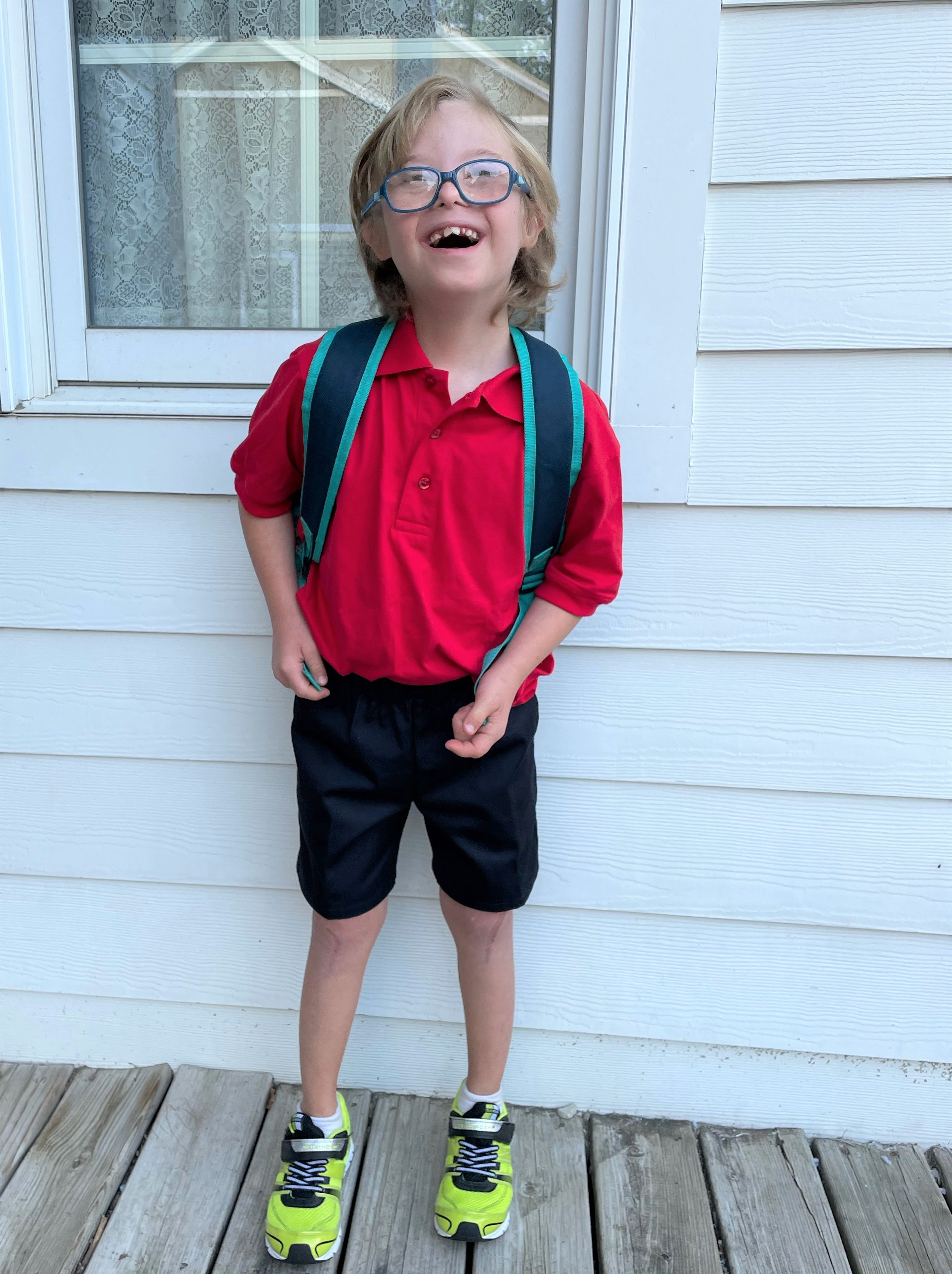 A young boy with glasses, blonde hair, and a big smile, wearing a red polo shirt, black shorts, and bright yellow sneakers, standing on a wooden deck outside a house with white siding and a window behind him. He has a blue and black backpack.