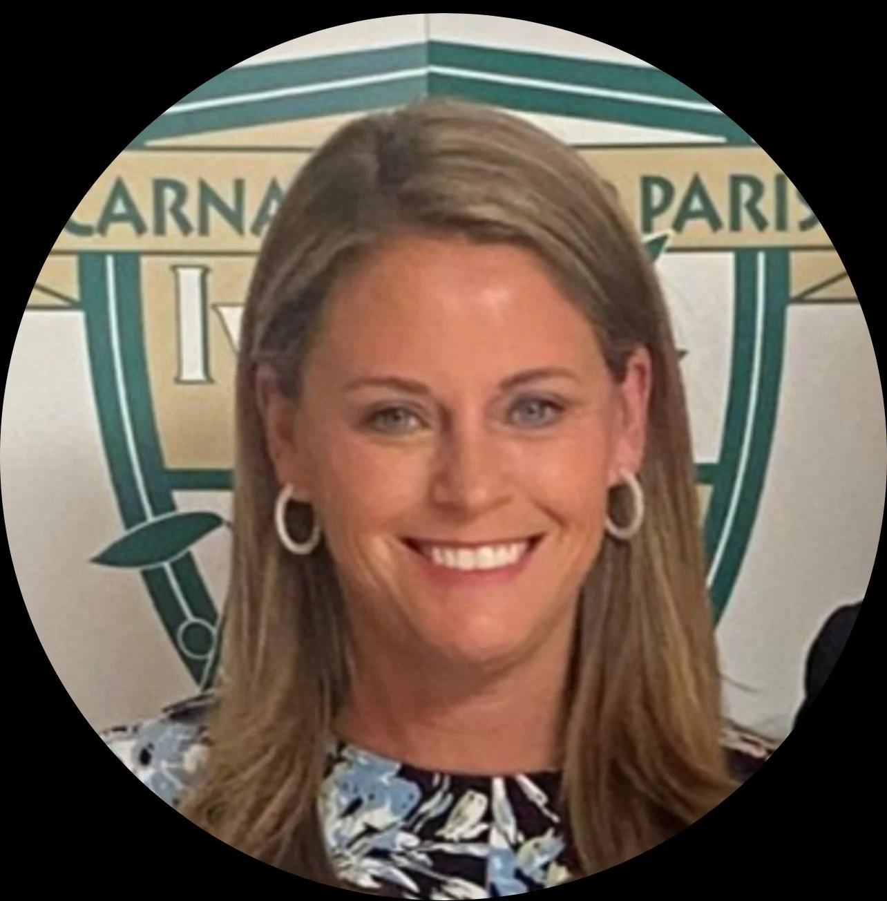 A woman with long blonde hair, wearing earrings and a patterned blouse, smiling in front of a logo with the words 'Larna, Paris' and a crest.