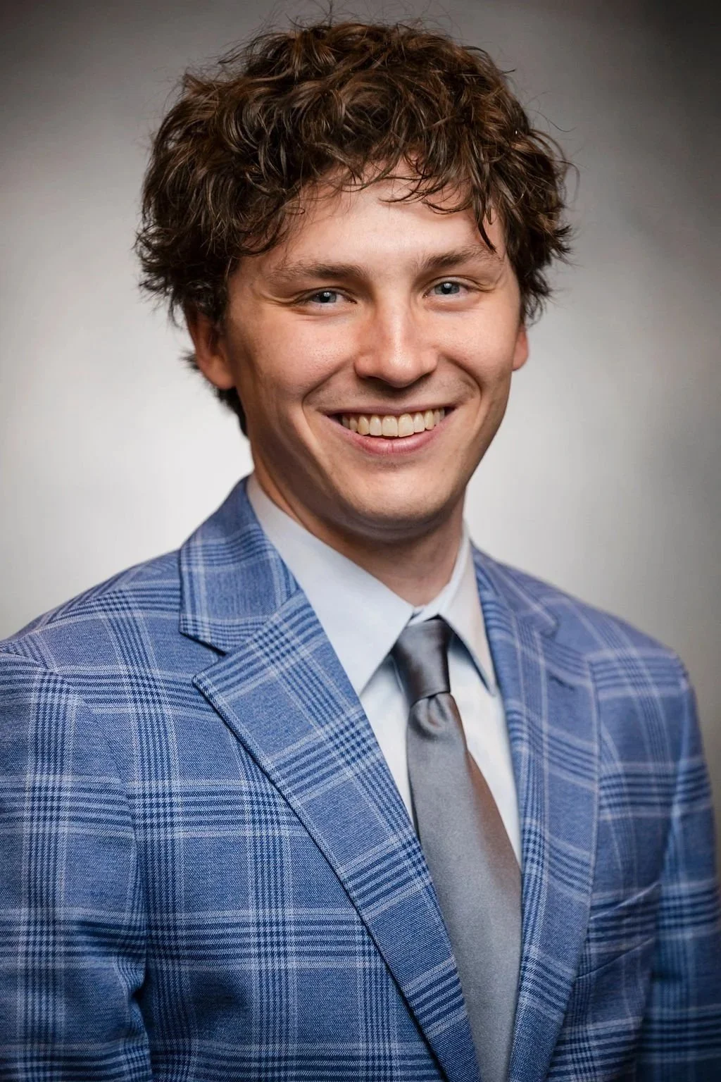 A young man with curly brown hair and blue eyes wearing a blue plaid suit, white shirt, and gray tie, smiling in a professional portrait.