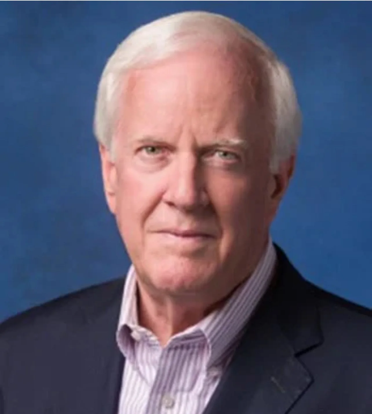 A portrait of an older man with light skin, silver hair, and blue eyes, dressed in a dark suit jacket and a striped white shirt, against a blue background.