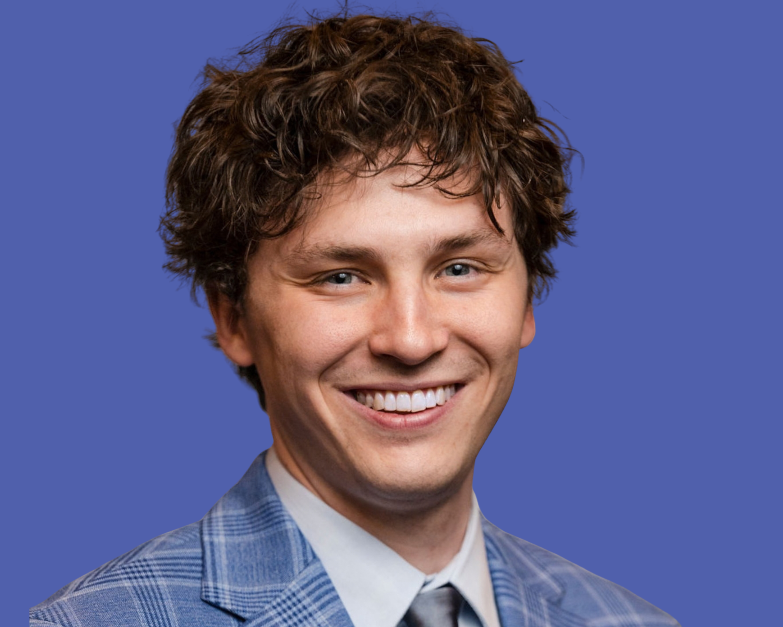 A young man with curly brown hair and blue eyes wearing a blue plaid suit, white shirt, and gray tie, smiling in a professional portrait.