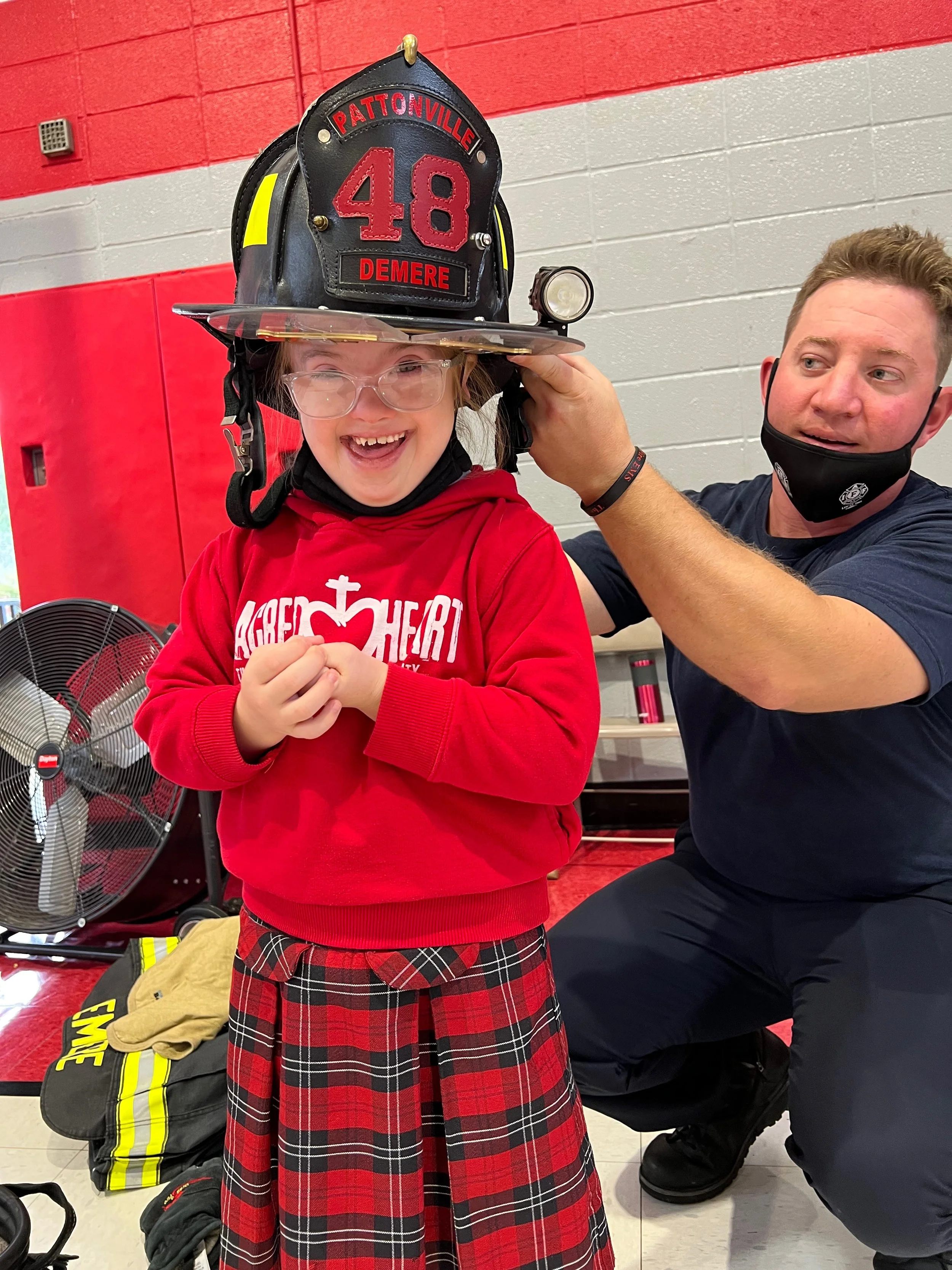A young girl wearing glasses and a red hoodie with a firefighter helmet on her head, smiling. An adult male firefighter assists her, adjusting the helmet. The helmet has the number 48 and the name Demere on it. In the background, there are firefighting gear and a fan.