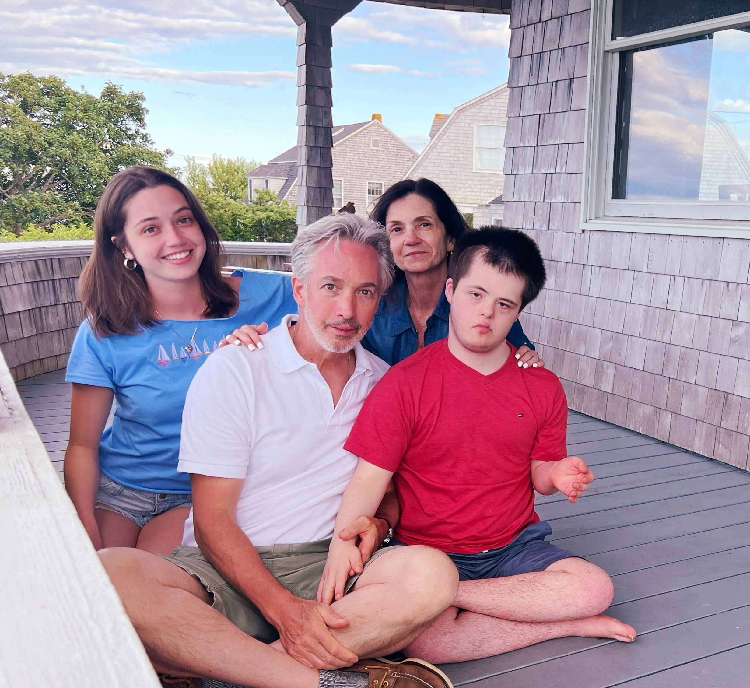 A family of five sitting on a porch. The group includes two women, two boys, and one older man. They are smiling and looking at the camera, with a blue sky and neighboring houses in the background.