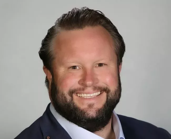 A man with a beard and mustache, smiling, wearing a suit jacket and shirt against a plain background.