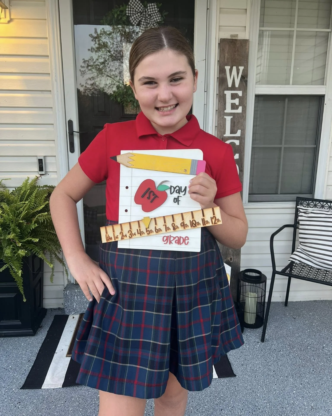 A girl in a red polo shirt and plaid skirt holding a sign about the "First Day of 1st Grade" standing outside a house with a welcome sign, a plant, and outdoor furniture.