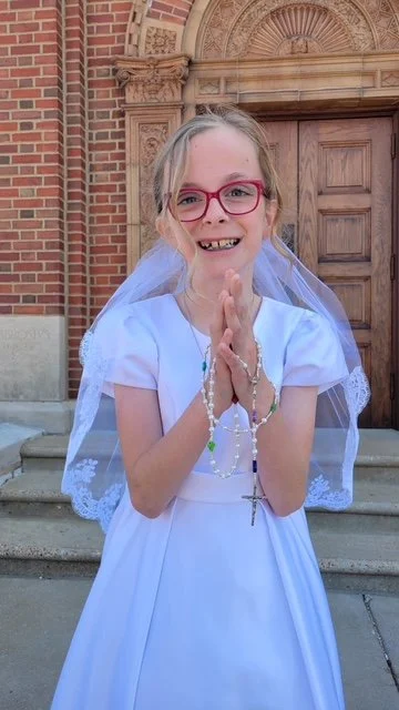 A young girl dressed in a white dress with a veil, holding rosary beads and standing in front of a brick church.