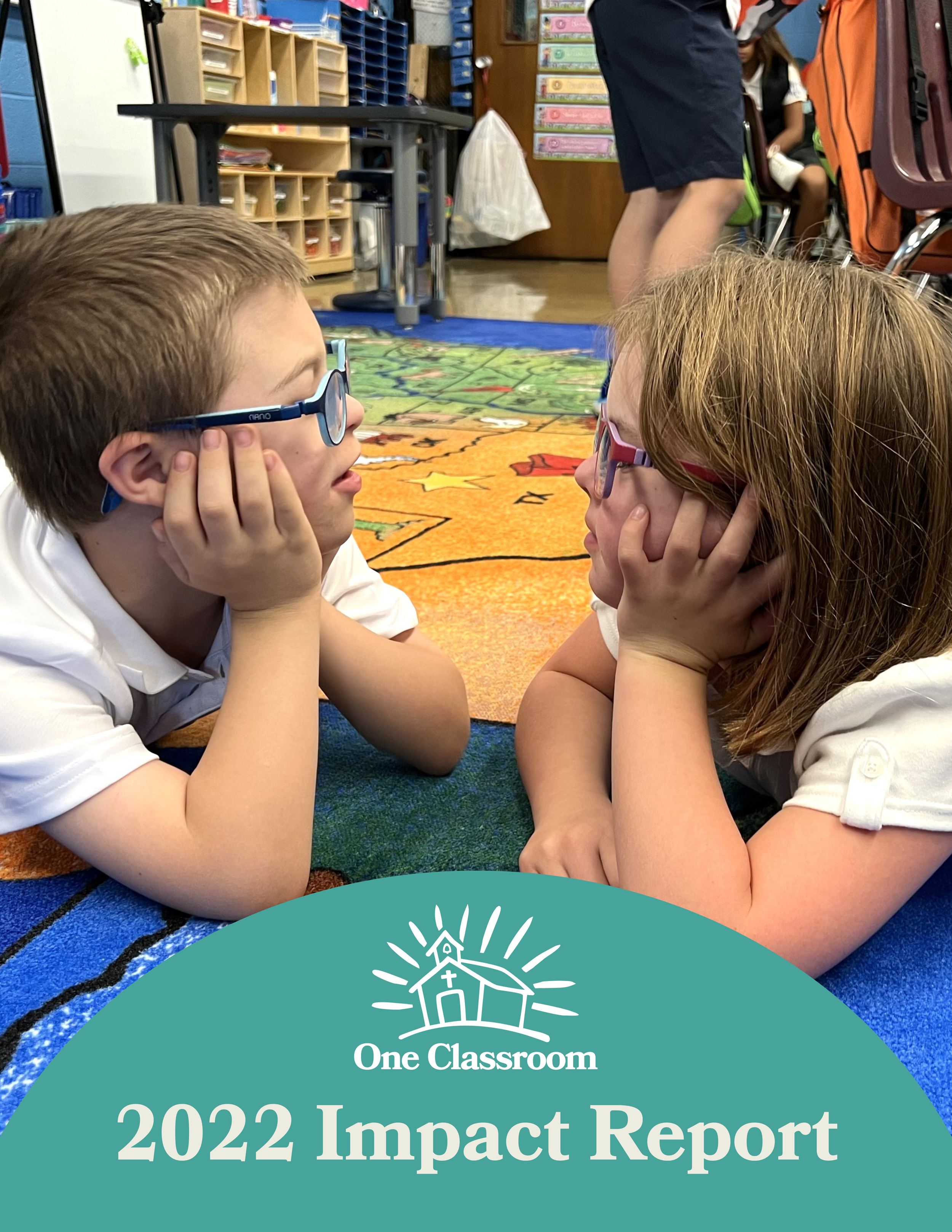 Two children, a boy and a girl, are lying on a colorful carpet in a classroom, facing each other and resting their chins on their hands, engaging in a conversation. They are wearing glasses and school uniforms. In the background, classroom shelves, a teacher, and other students are visible.