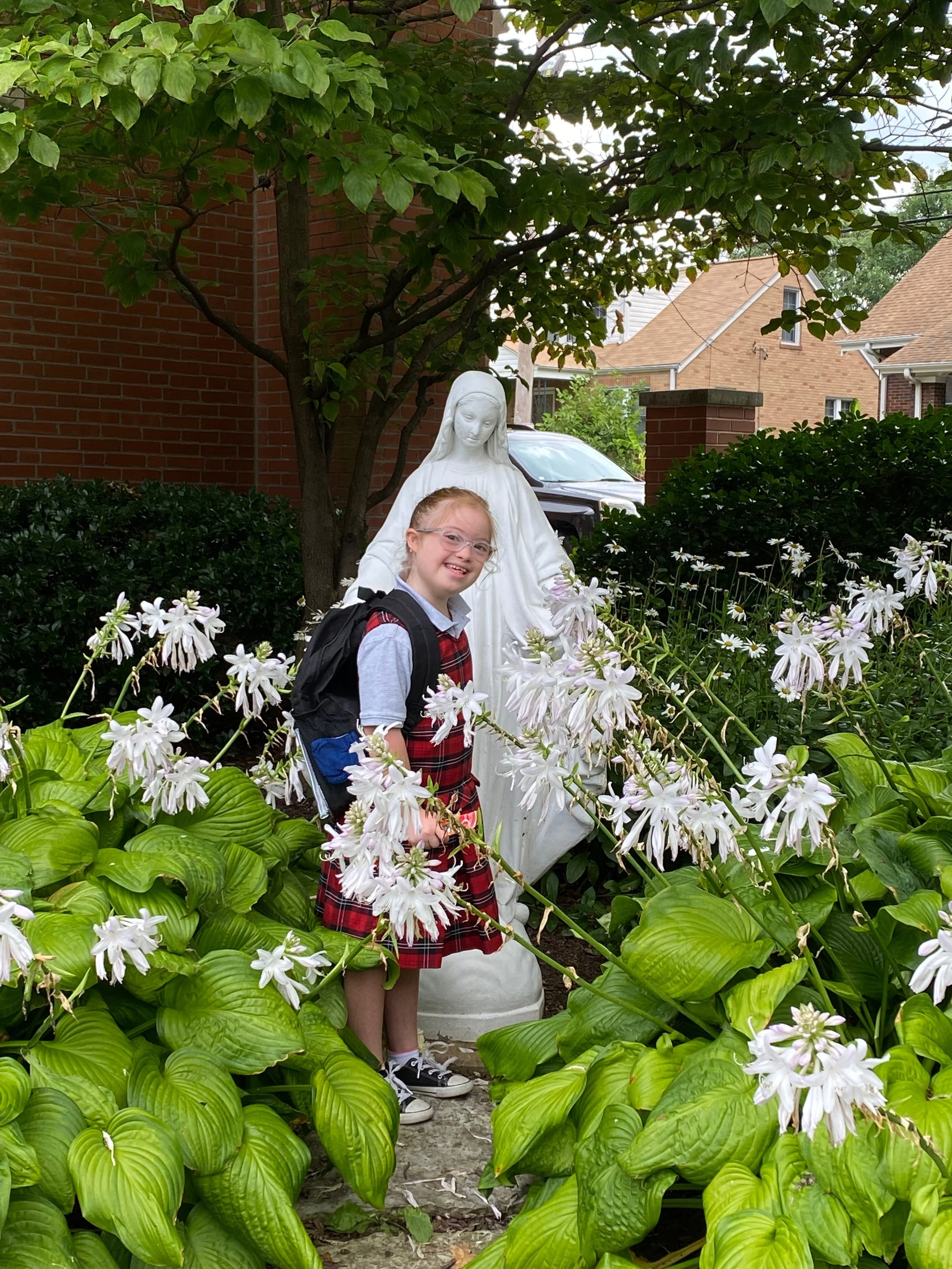A young girl with glasses, a backpack, and a red plaid dress standing among white flowers, with a white statue of Mary behind her and green foliage around.