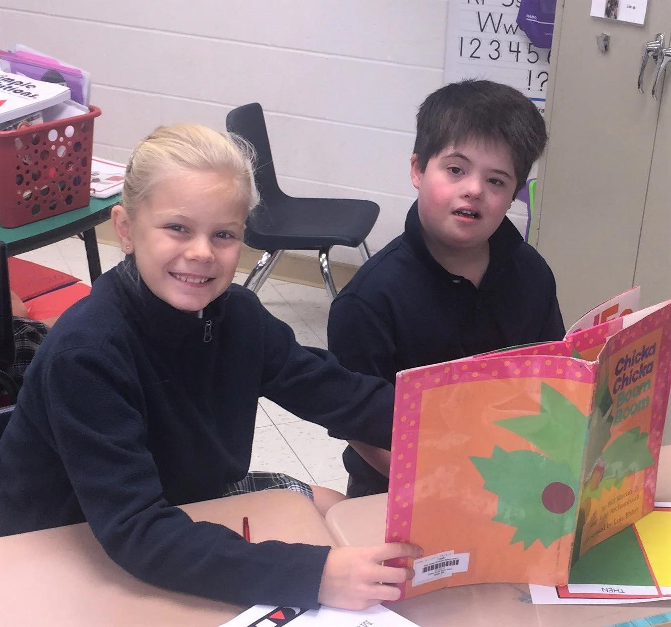 Two children, a girl with blonde hair and a boy with dark hair, sitting at a table in a classroom, looking at the camera. They are reading a colorful book titled 'Chicka Chicka Boom Boom'.