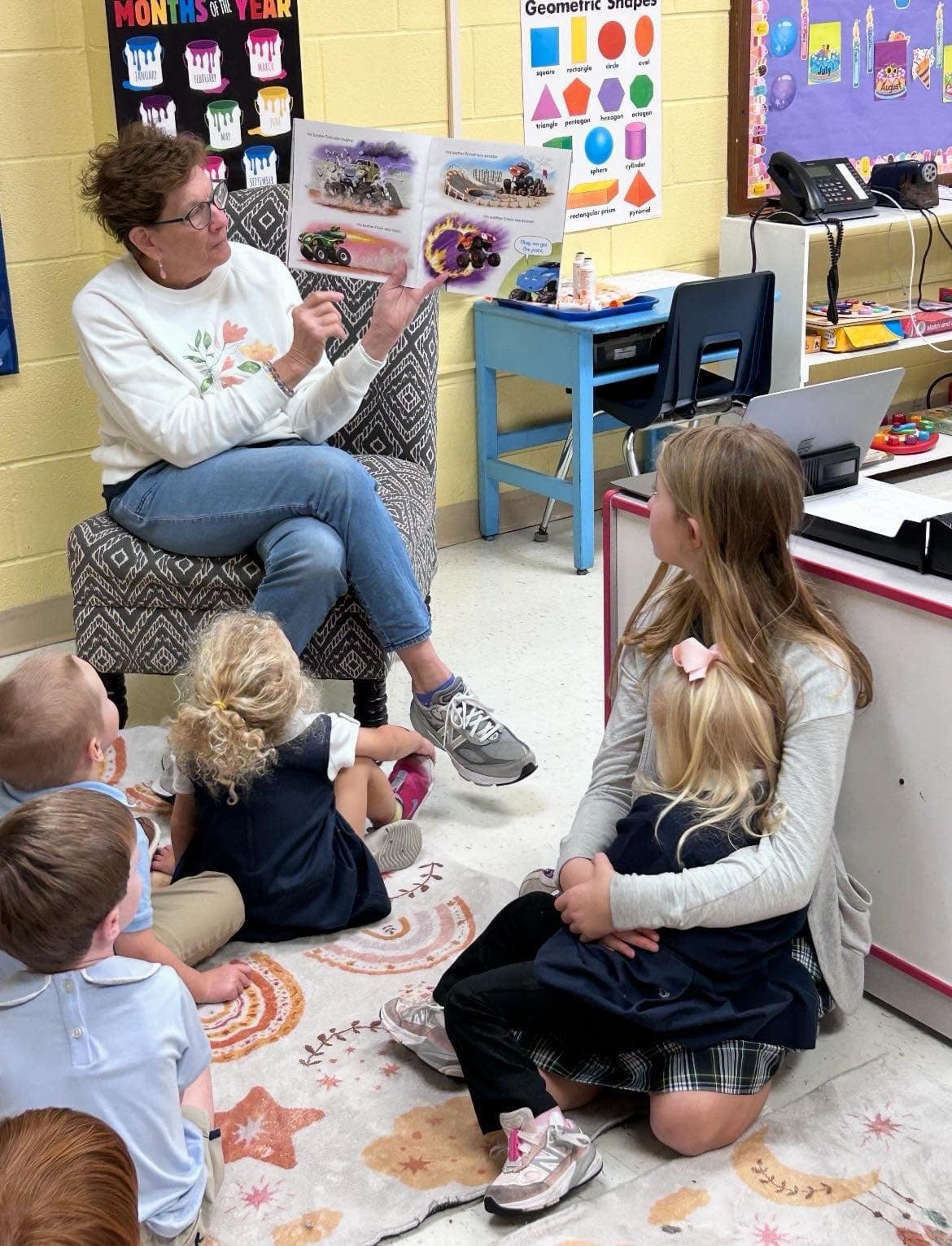 Teacher reading a picture book to a group of young children sitting on a colorful rug in a classroom.