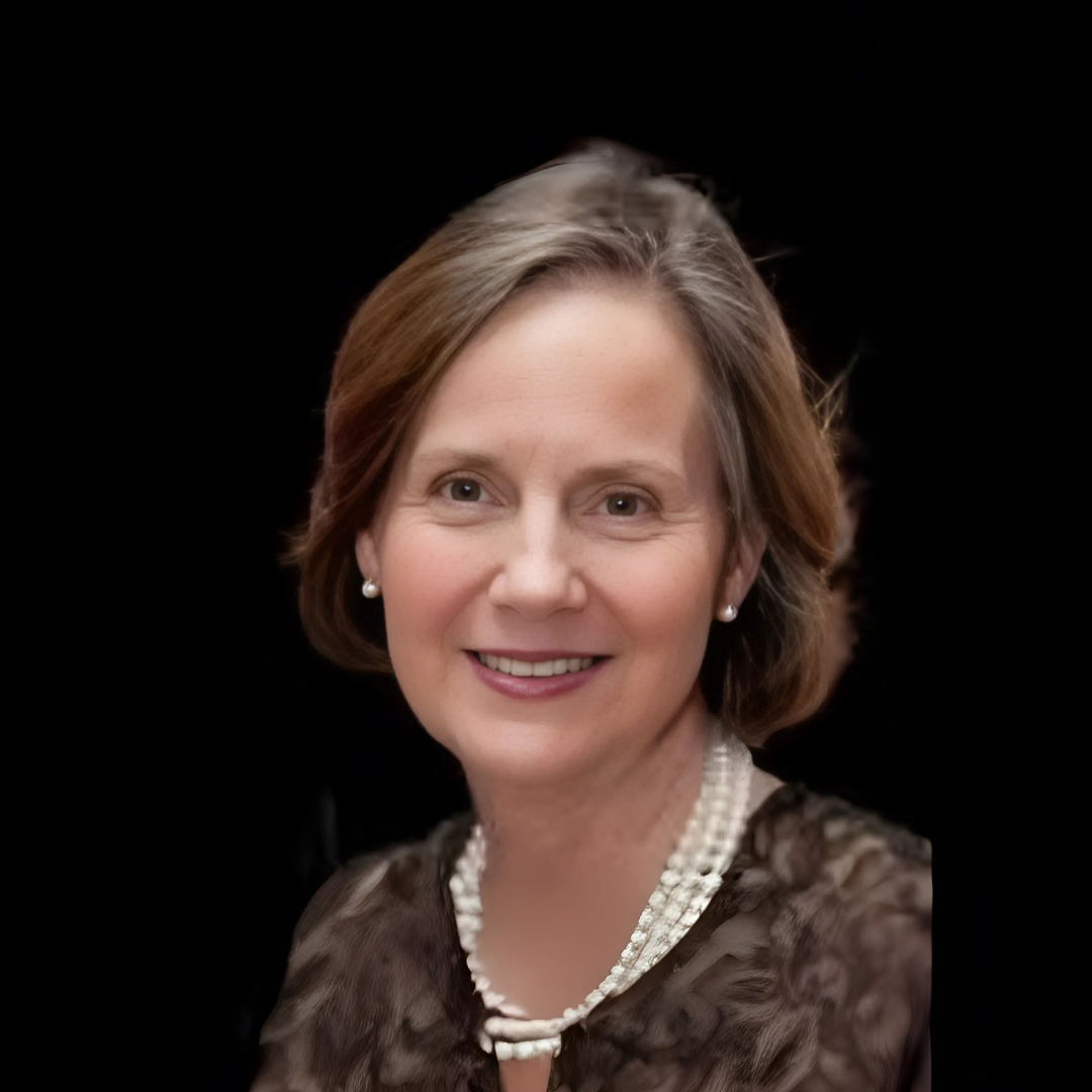 Smiling middle-aged woman with short brown hair, pearl earrings, a pearl necklace, and a patterned top, against a black background.