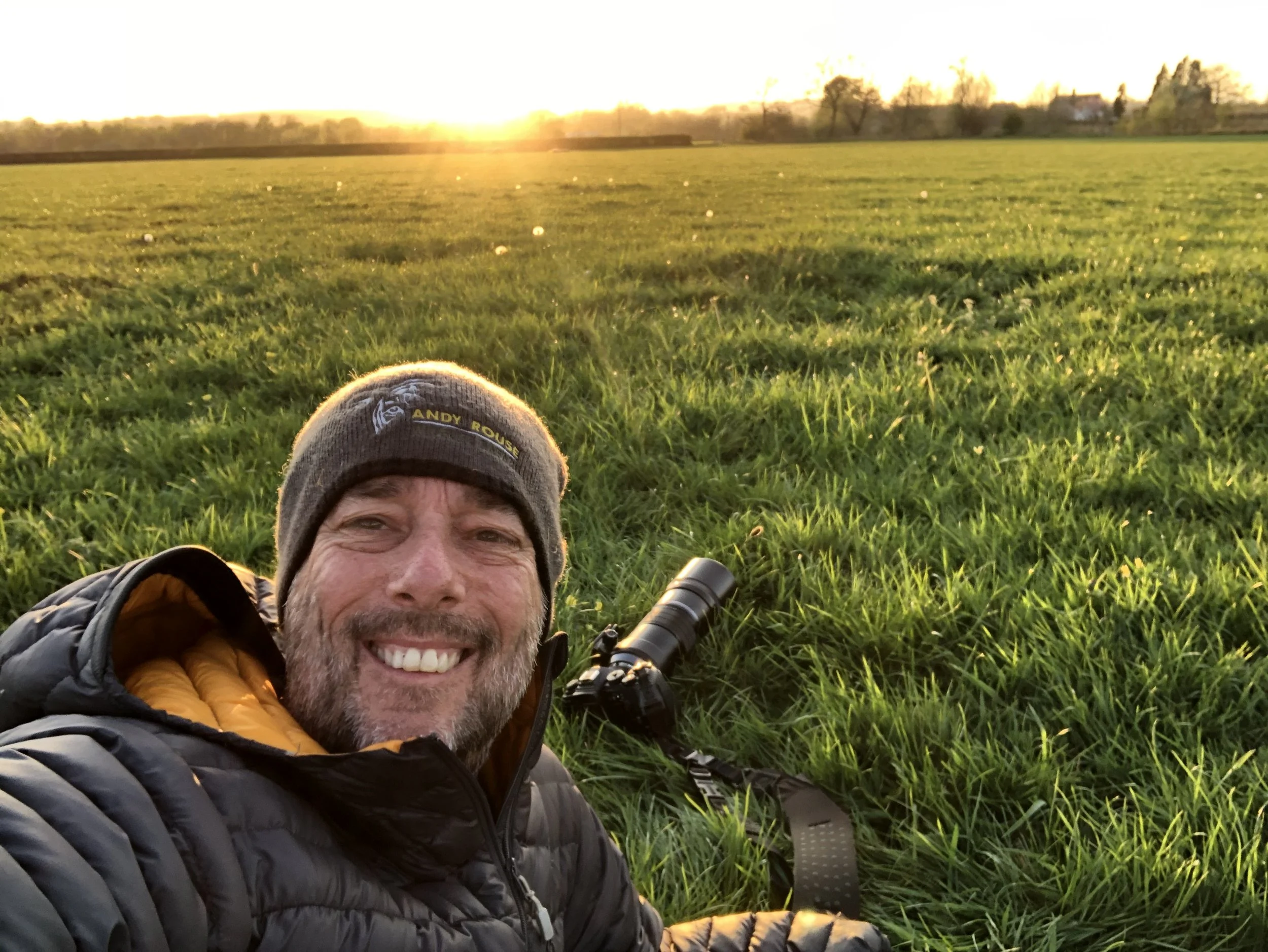 Smiling man taking a selfie in a grassy field at sunset, with a camera lying on the grass nearby.