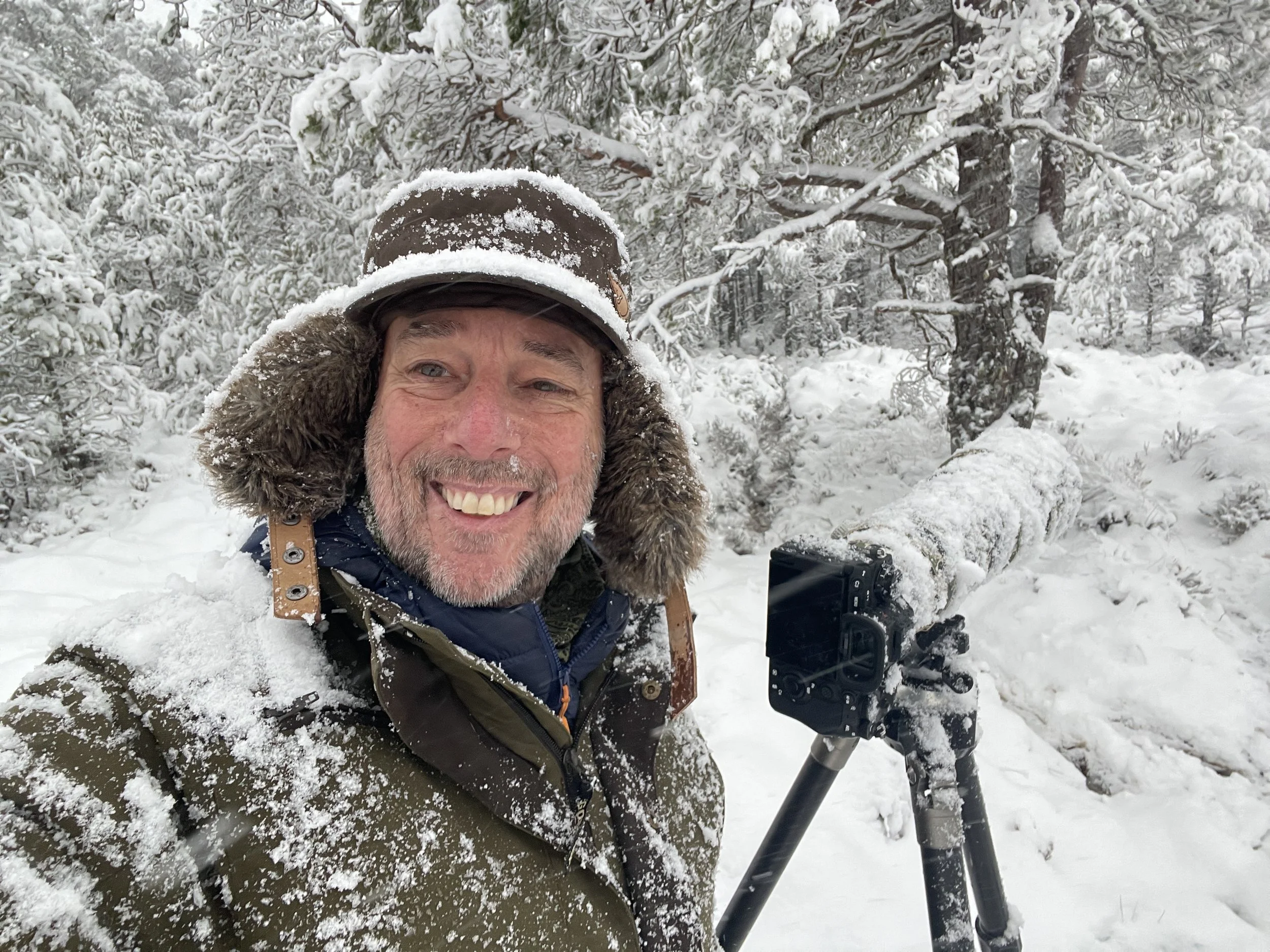 Man in winter gear smiling in snowy forest with camera on tripod.