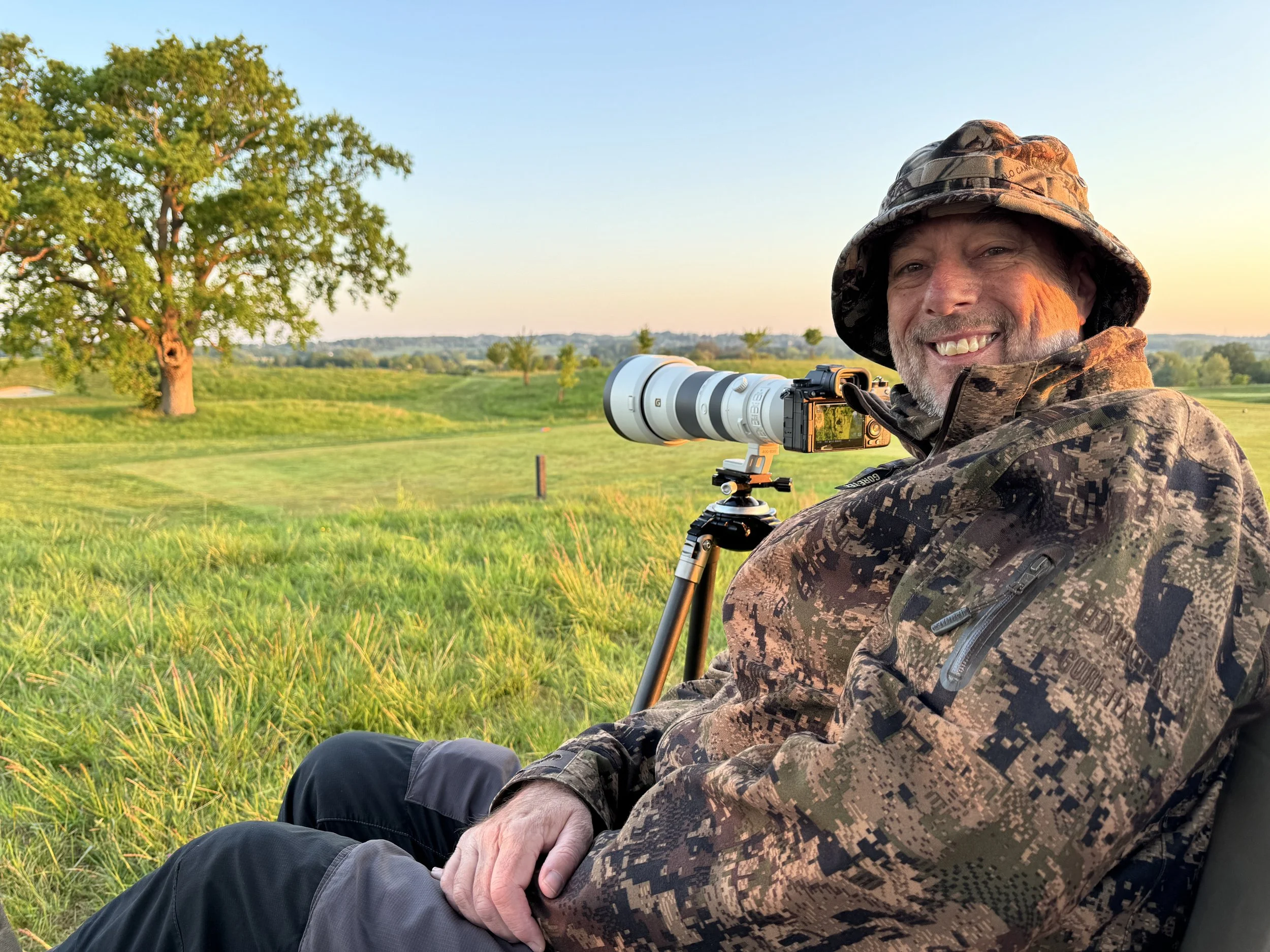 A man in camouflage clothing and a hat sitting outdoors with a telephoto camera lens on a tripod, smiling at the camera during sunset, with a large tree and open field in the background.