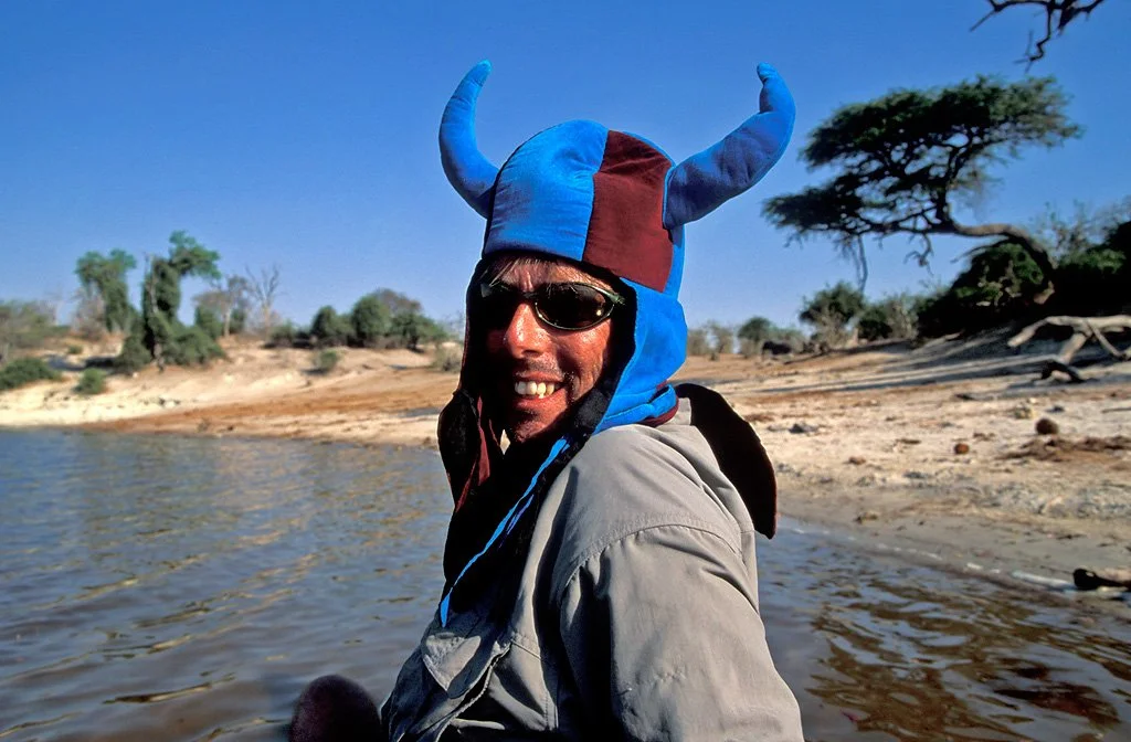 Person wearing a blue and brown Viking hat with horns, sunglasses, and a gray jacket, standing near a river with trees and a sandy shoreline in the background.