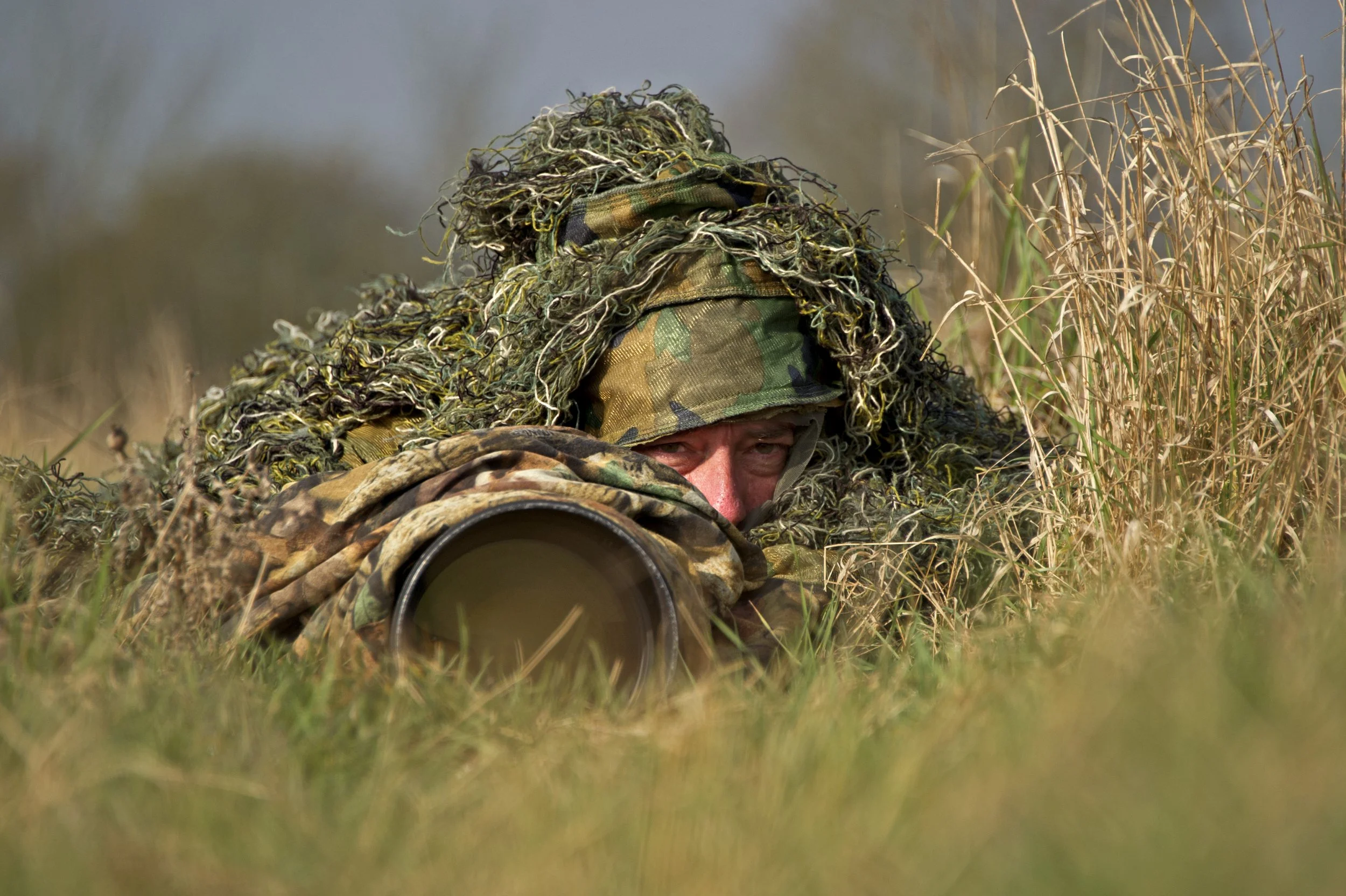 A soldier in camouflage gear lying prone in tall grass, aiming a rifle through a scope, with a ghillie suit blending into the surroundings.