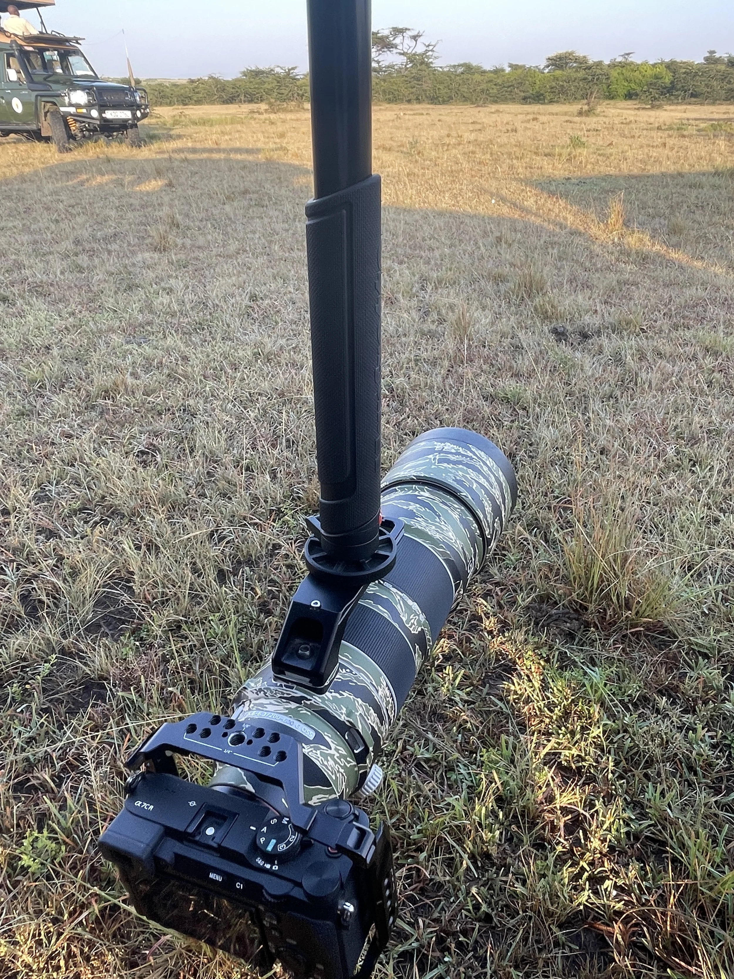 Camera mounted on a tripod with a camouflage pattern, set up on a grassy field in a savanna landscape with two safari vehicles in the background.