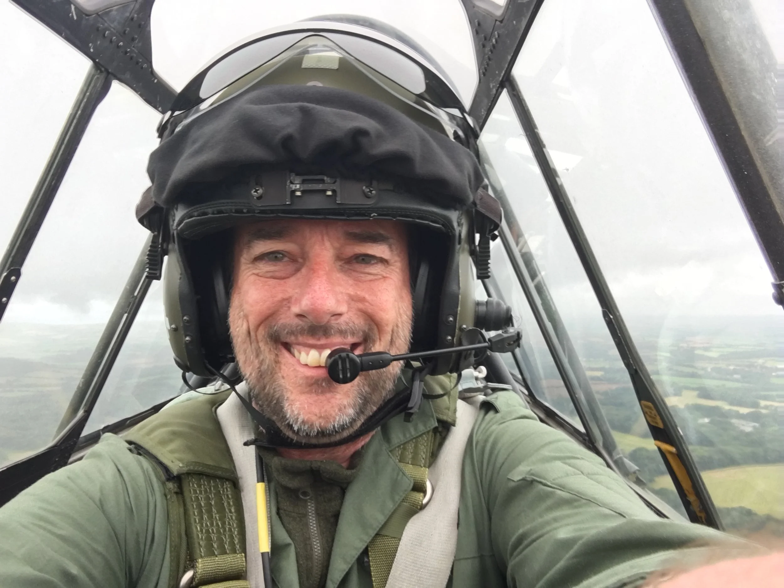 A man smiling in the cockpit of a small aircraft, wearing a pilot helmet and flight gear.