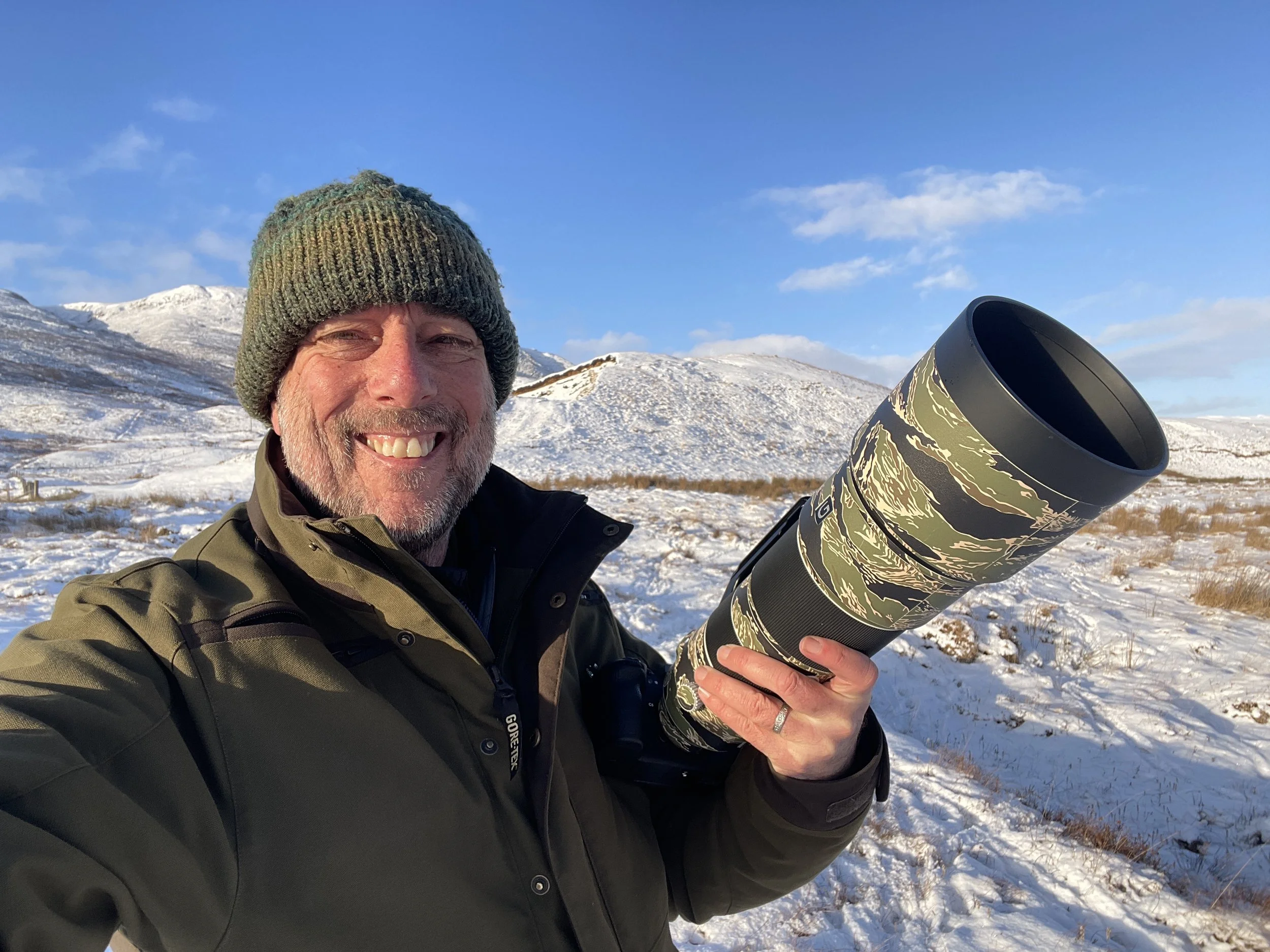 Smiling man in outdoor winter clothing holding a camera lens in snow-covered landscape with hills and blue sky