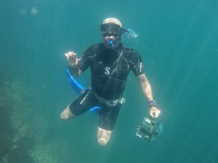 A man scuba diving underwater, wearing a black wetsuit, mask, and snorkel, holding an underwater camera, and making the peace sign with his right hand.